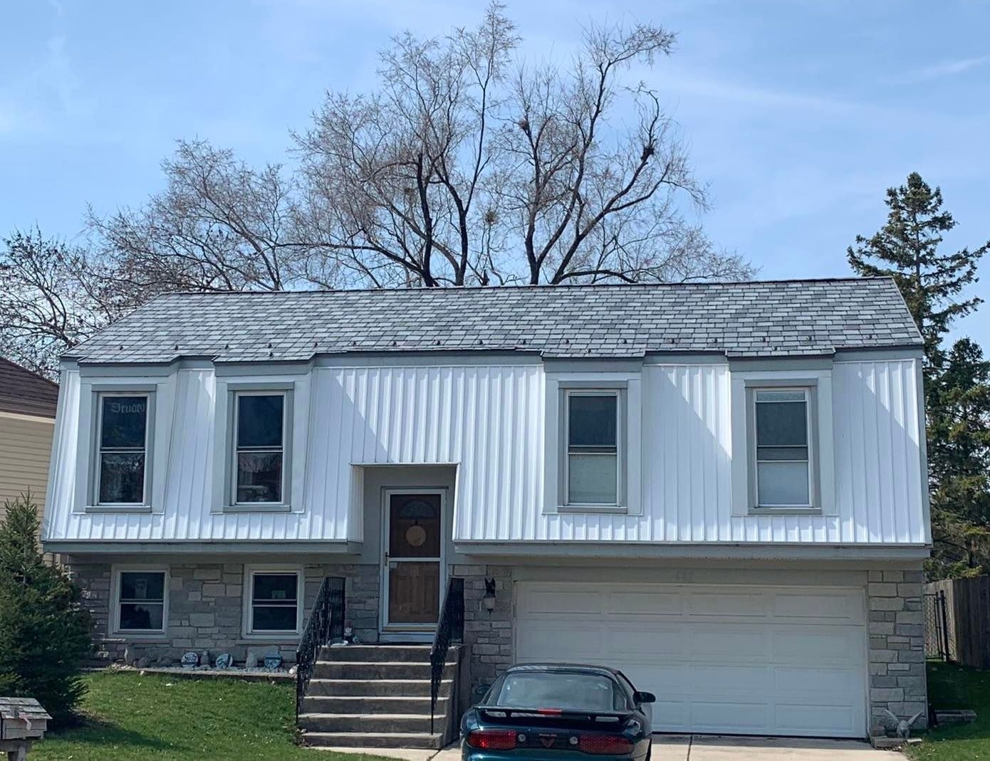 Two-story house with light siding and grey roof. A car parked in front of a garage. A large tree behind the house.