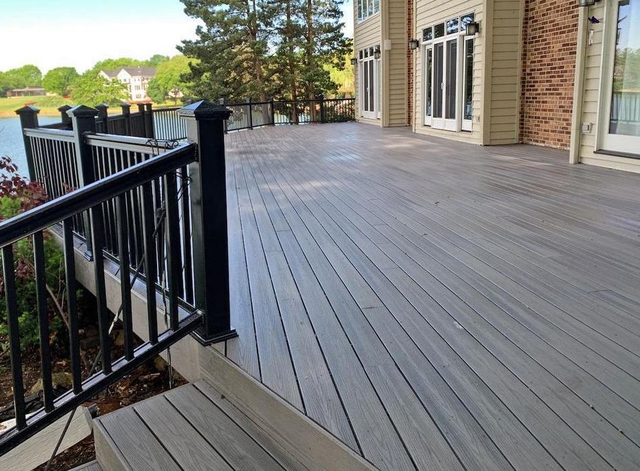 A large, grey composite deck with black railings overlooking a body of water next to a suburban house.