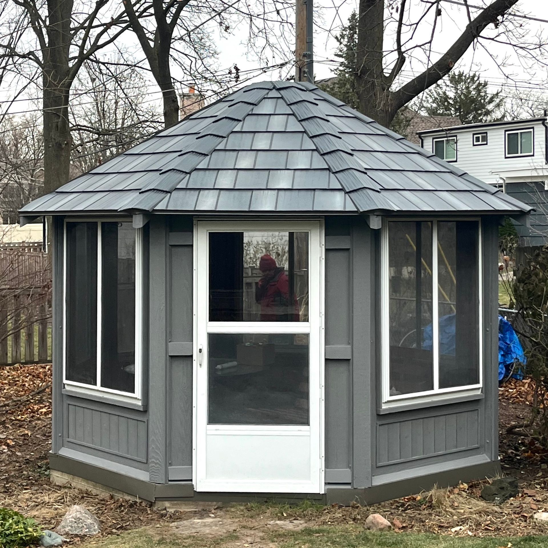A gray, hexagonal gazebo with a shingled roof, white door, and windowed panels in a backyard setting.