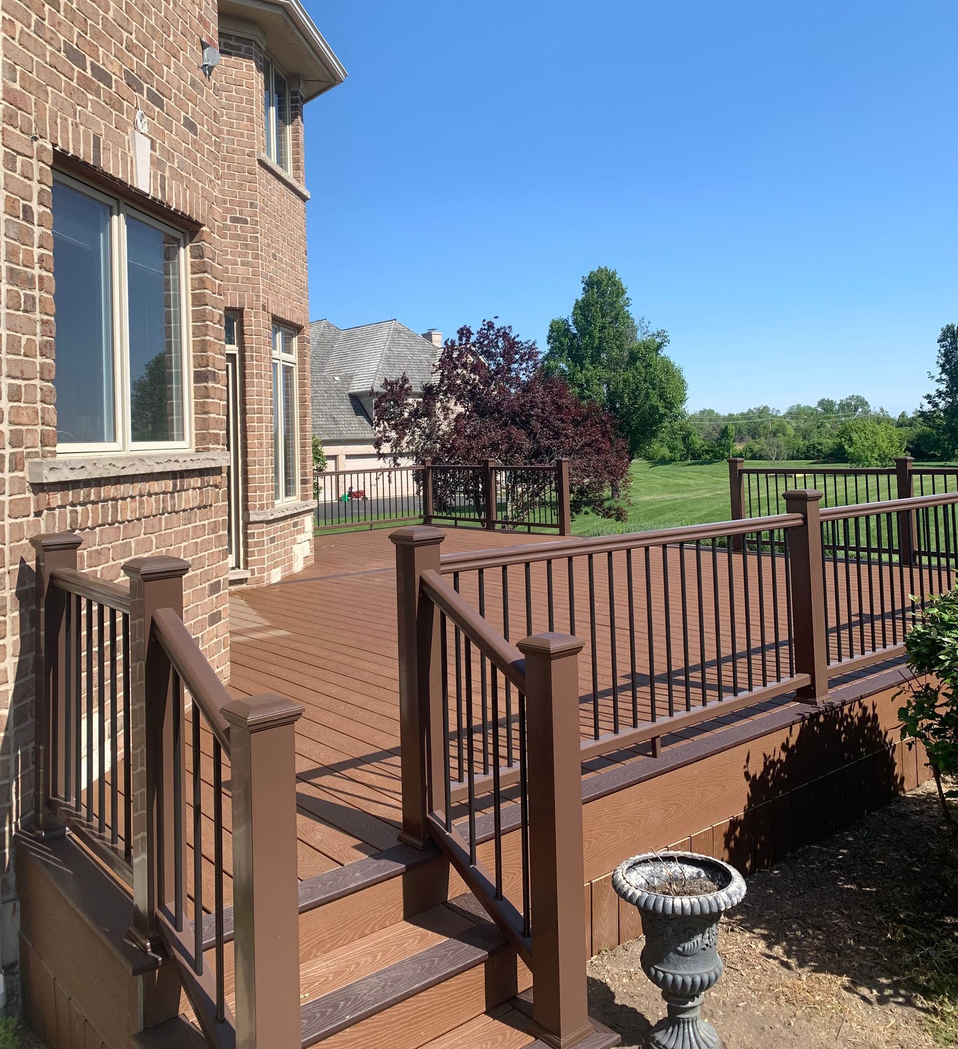 A brown composite deck attached to a brick house, featuring dark railing, stairs, and a view of a grassy yard.