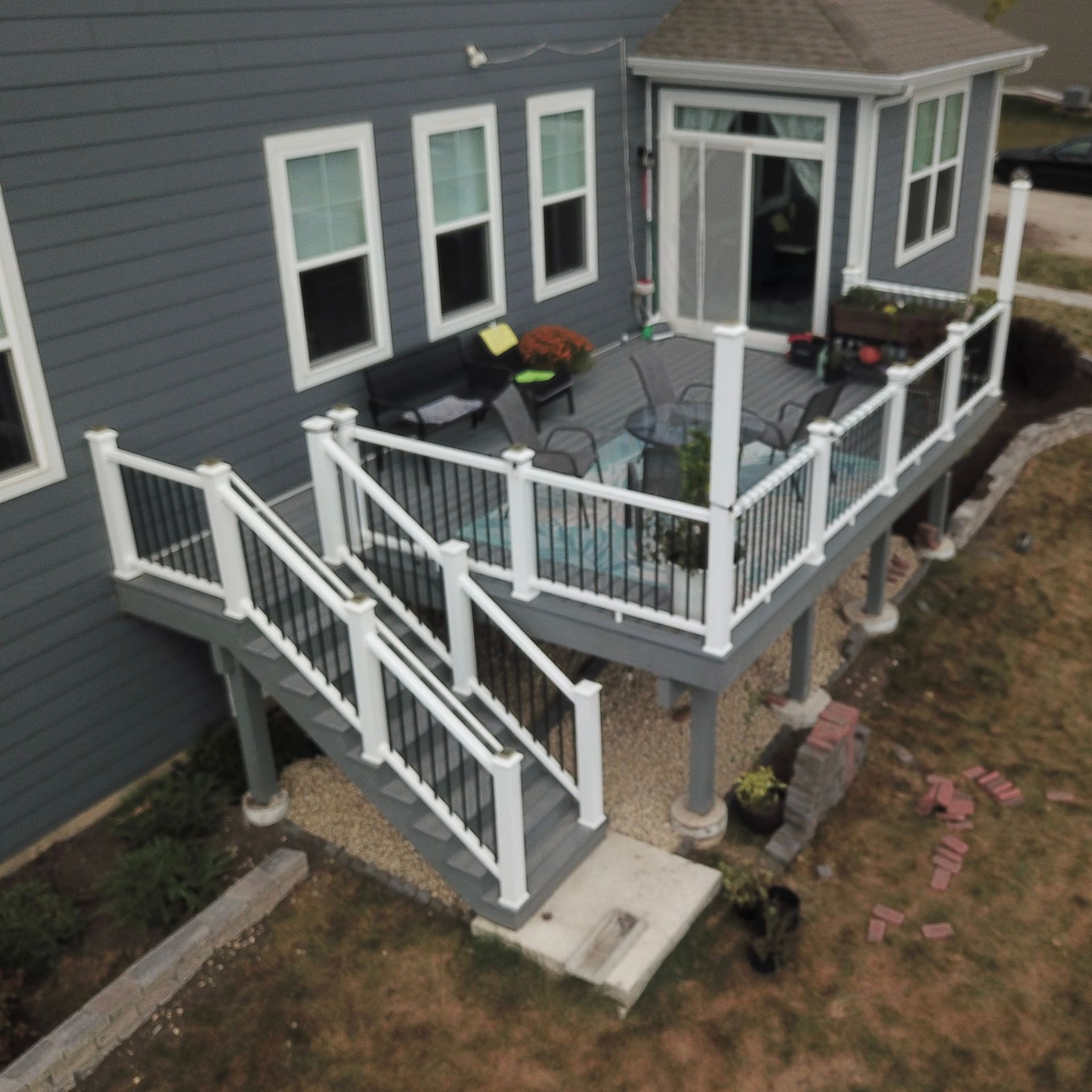 A gray house with a raised wooden deck, white railings, and a staircase leading down to a small concrete patio.