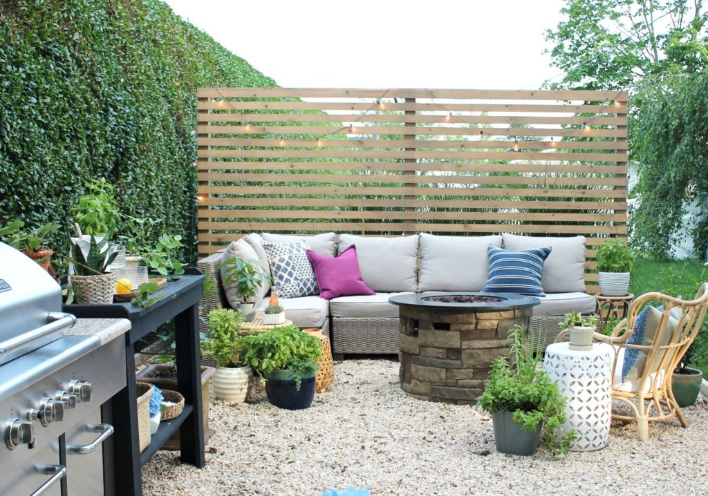 A patio with a beige sectional sofa, fire pit, grill, and wood slat privacy screen on a gravel surface.