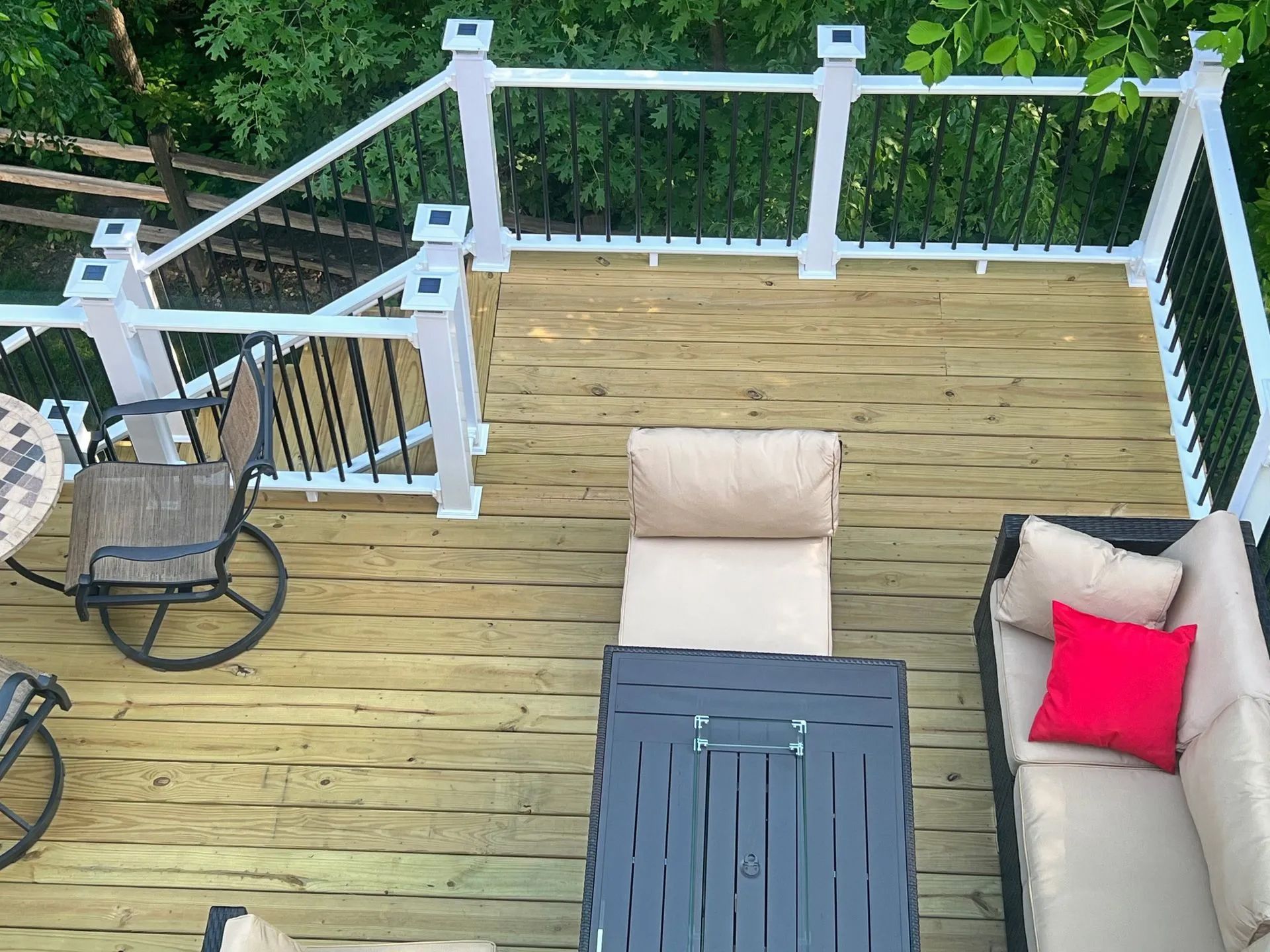 A high-angle view of a wooden deck featuring a patio set with tan cushions, a red throw pillow, and white railings.
