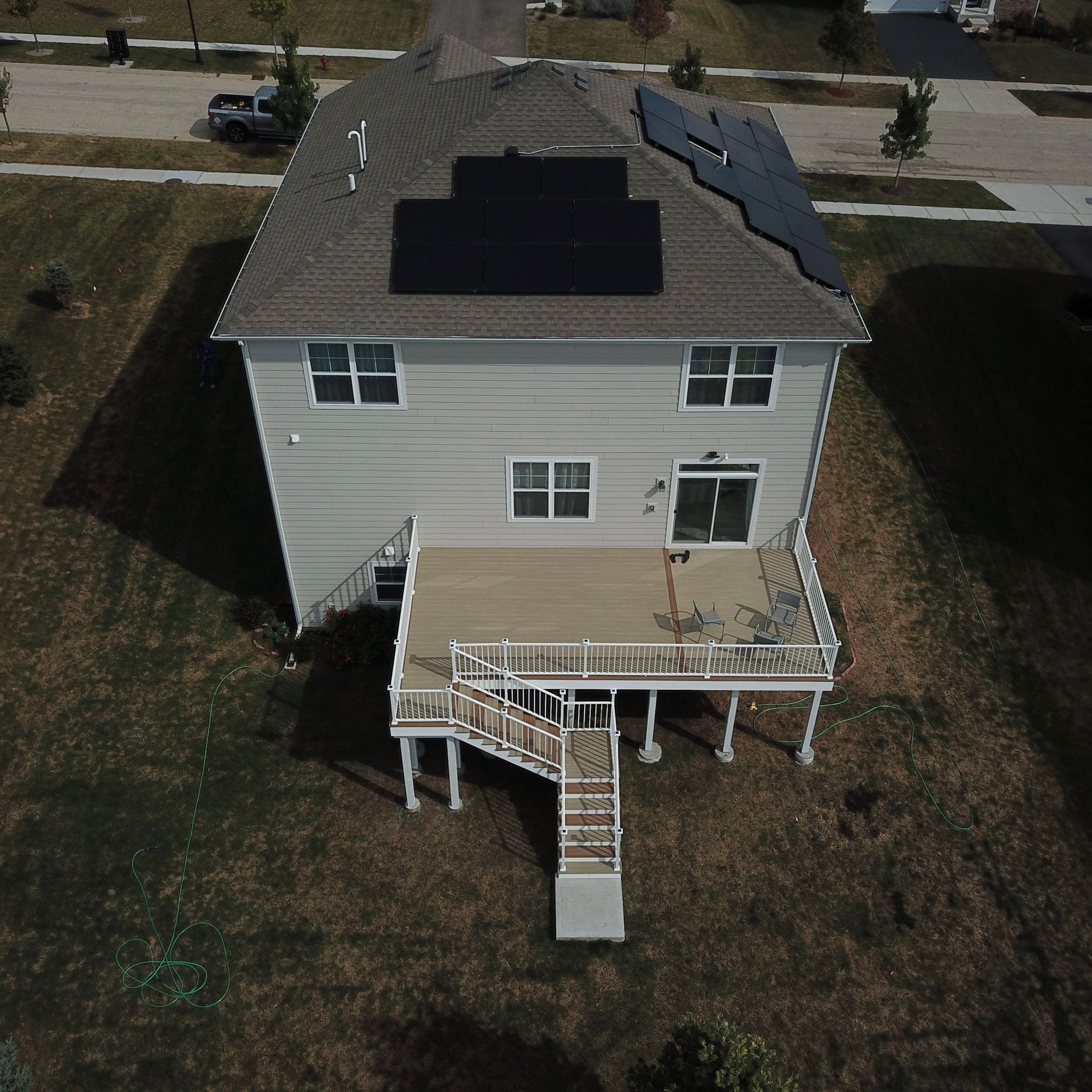 An aerial view of a suburban house with solar panels on the roof and a raised wooden deck with stairs in the backyard.