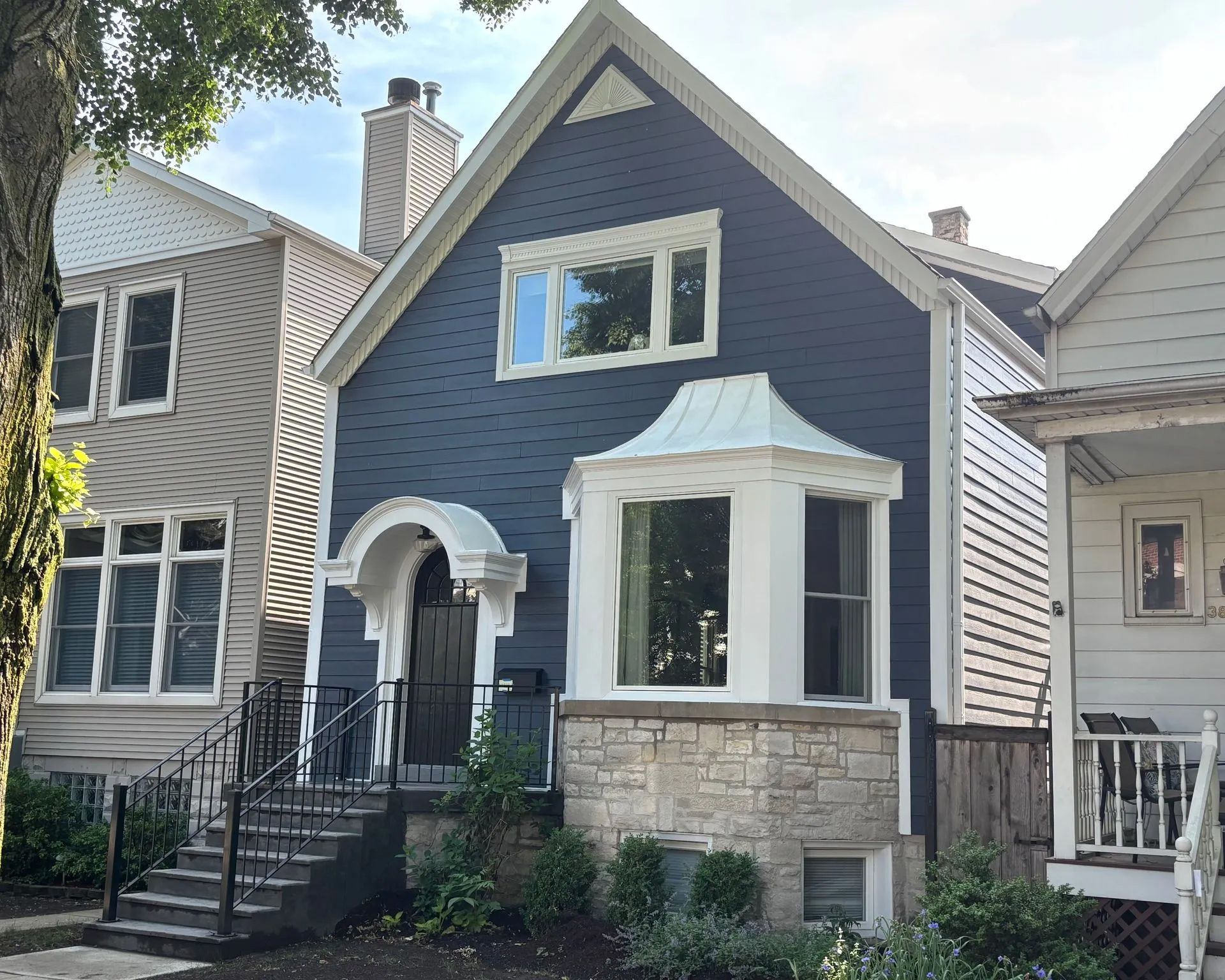 A blue house with a white bay window and arched entryway, flanked by two other houses on a tree-lined street.
