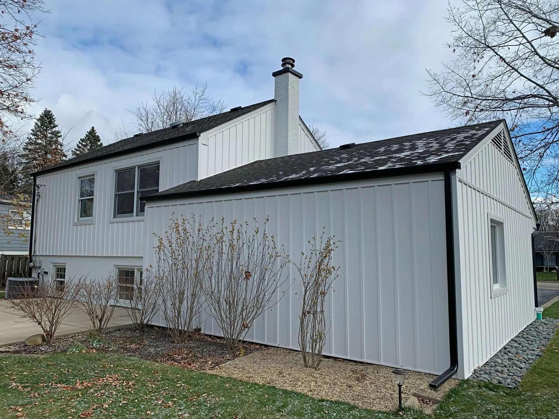 A white, single-story house with board-and-batten siding, a black shingled roof, and a stone chimney under a cloudy sky.
