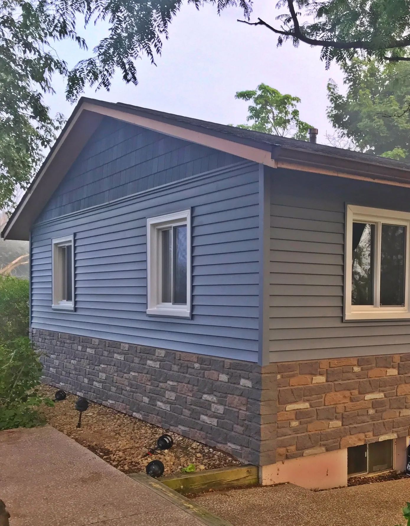 A house exterior with blue horizontal siding, a stone veneer base, and white-framed windows, set on a gravel lot.