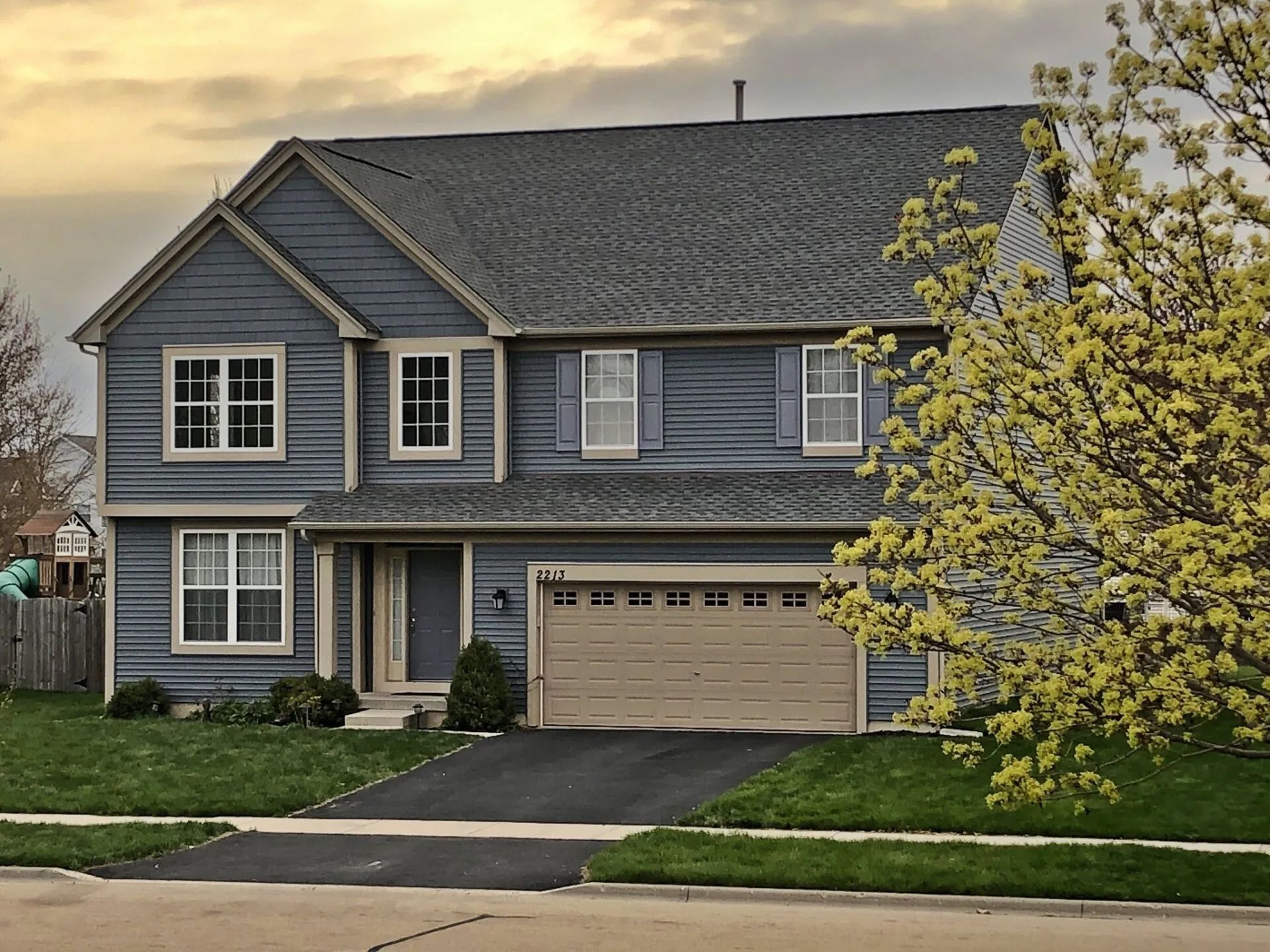 A two-story blue house with a beige garage, gray roof, and a flowering tree in the front yard at sunset.