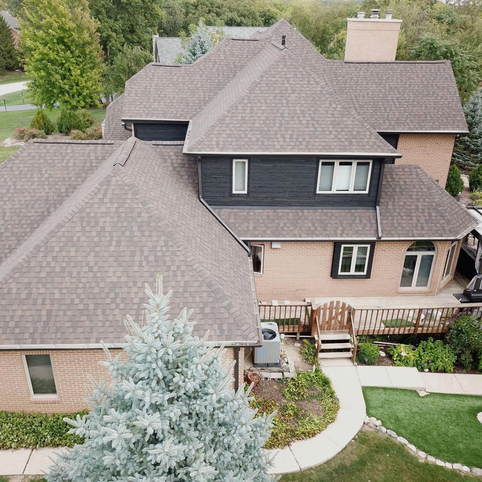 An aerial view of a two-story home with a brown shingled roof, tan brick, and dark siding, featuring a deck and yard.