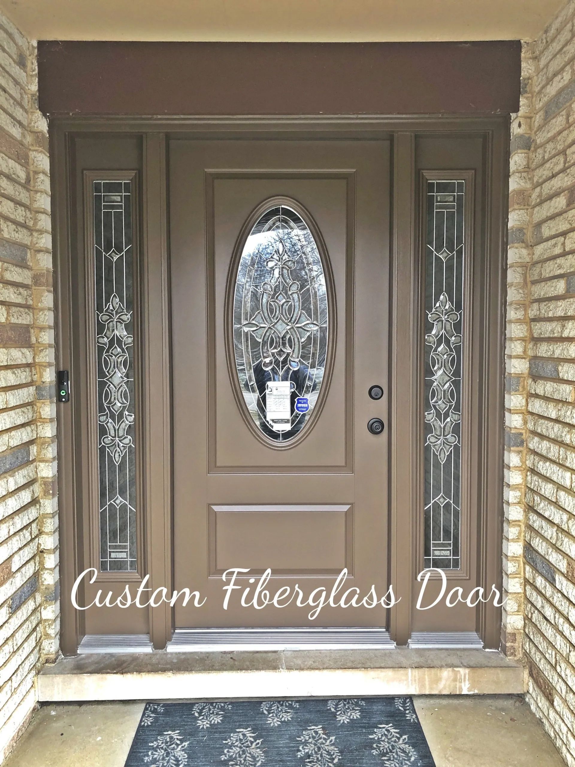 A brown custom fiberglass front door with an oval decorative glass insert and matching side lights, set in a brick frame.