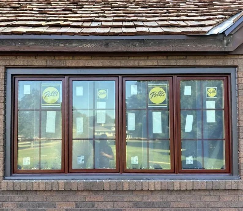 A four-pane brown-framed Pella window with grilles, installed in a brick wall below a cedar shake roof.