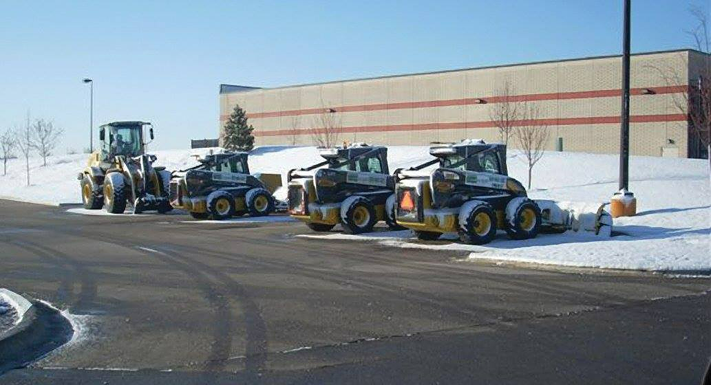 A row of snow plows is parked in a parking lot.