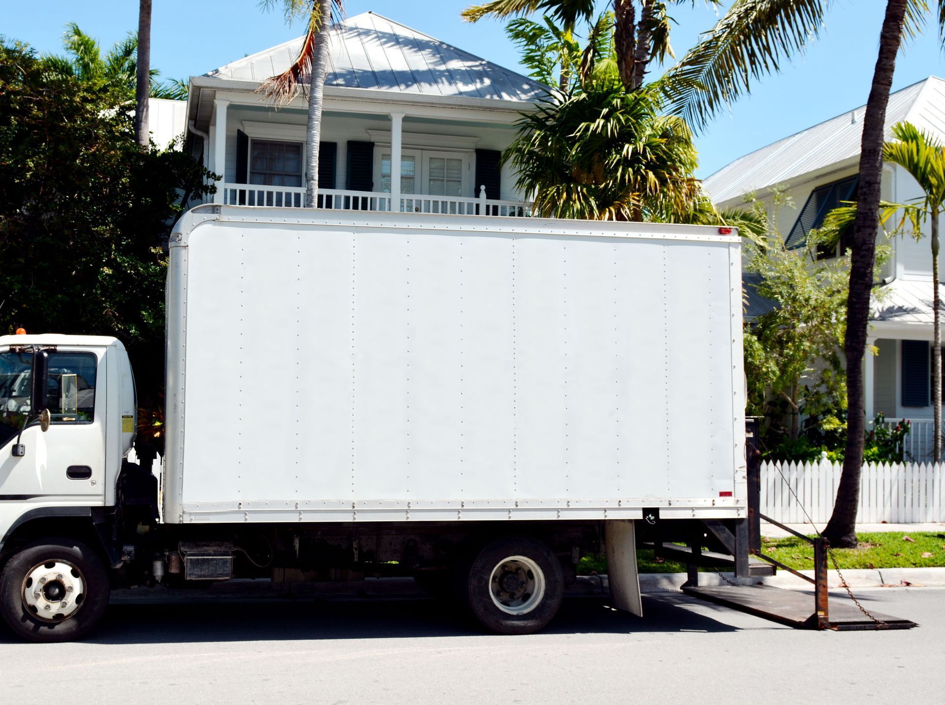 White box truck parked on a sunny street in front of a house with palm trees