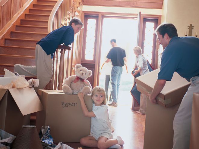 Family moving boxes in a bright hallway, with a child sitting in an open box near the stairs.