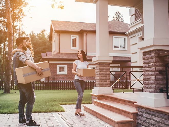 Two people carrying moving boxes outside a house at sunset