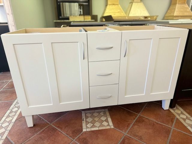 White bathroom vanity cabinet with three drawers and two doors, on a patterned tile floor.