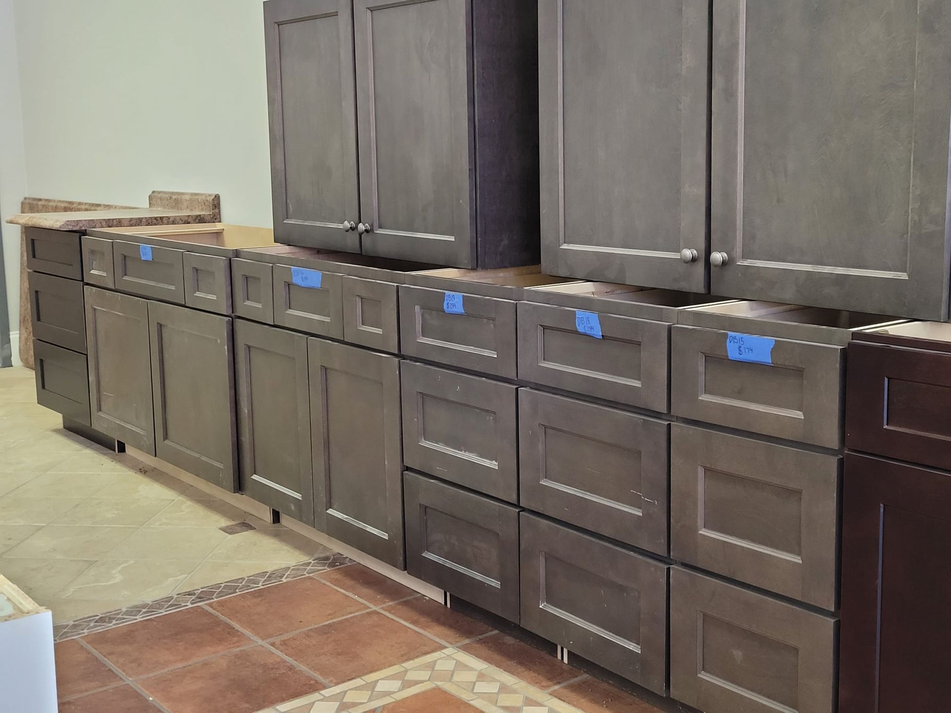 Kitchen cabinets, gray-brown, being installed. Base and wall cabinets visible. Brown tile floor.