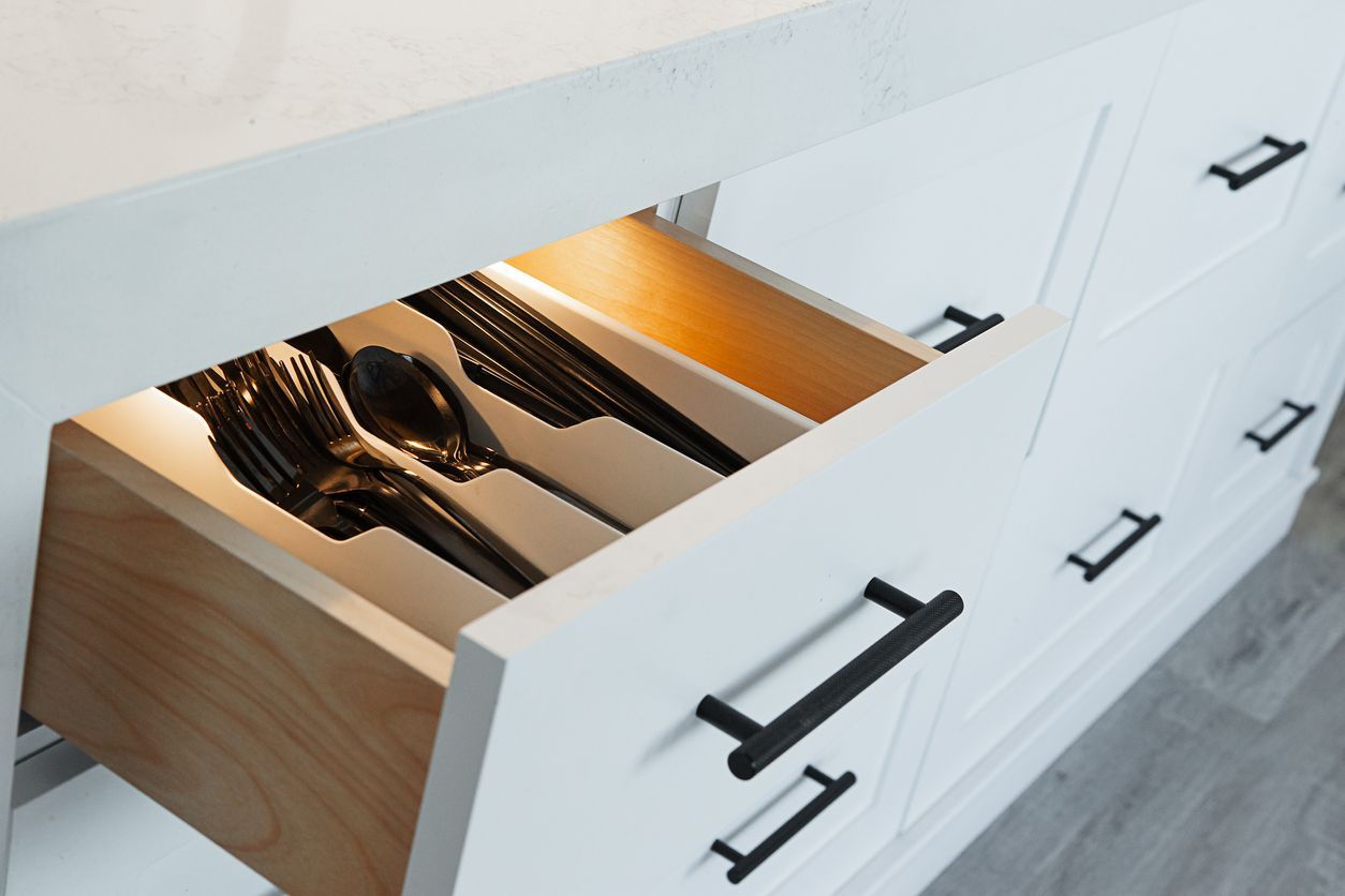 Open kitchen drawer with silverware. White cabinet with black handles, wooden drawer illuminated.