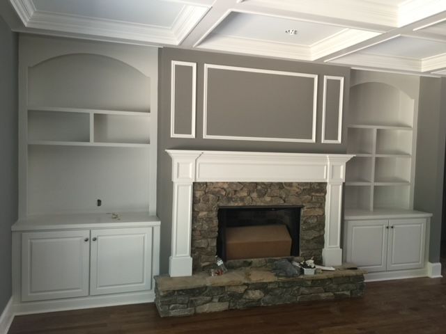 Built-in white bookshelves flank a fireplace with stone surround. Gray wall, white trim, and a coffered ceiling.