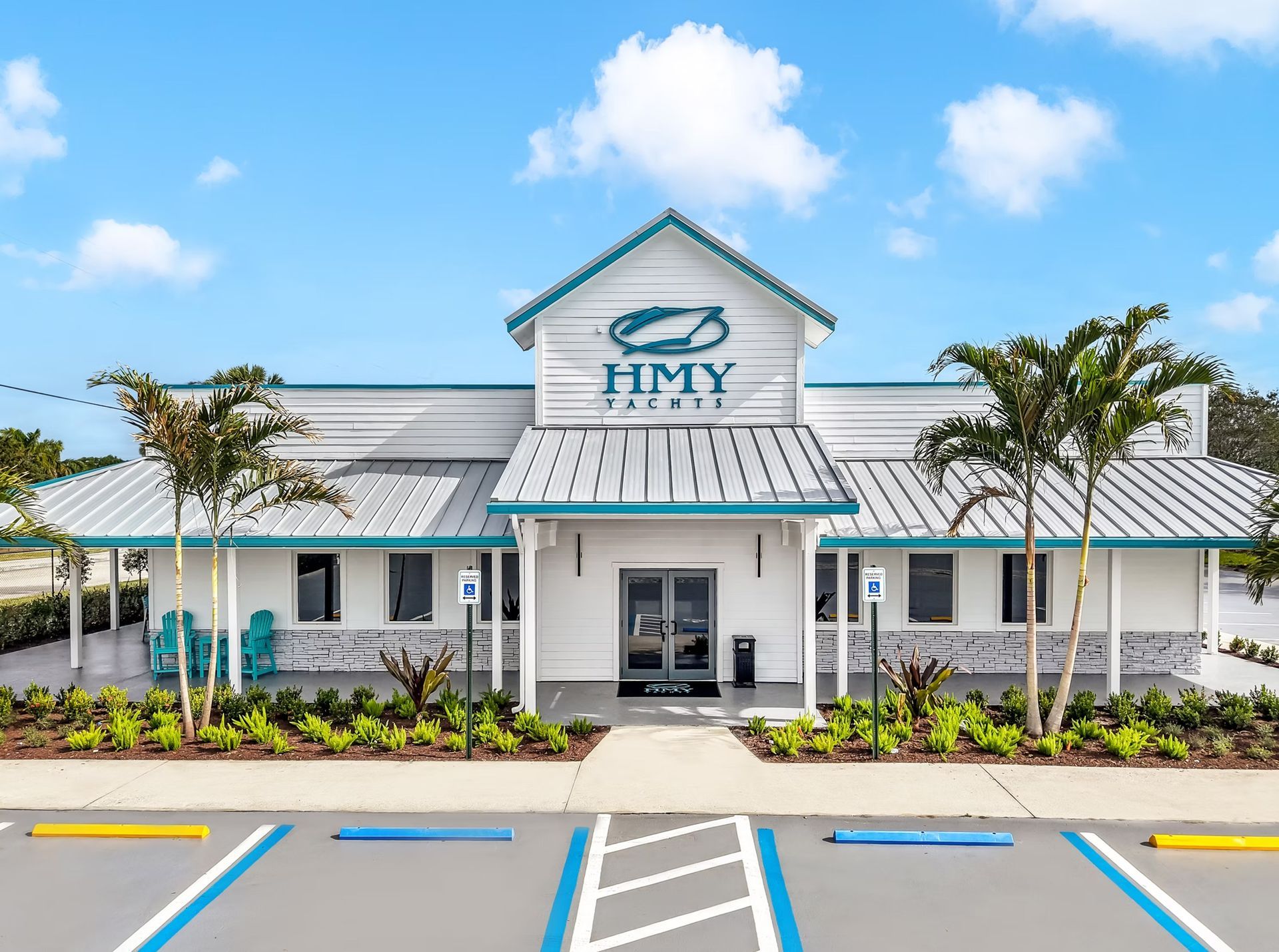 White building with teal trim and “HMY” sign, front entrance, parking spaces, and blue sky