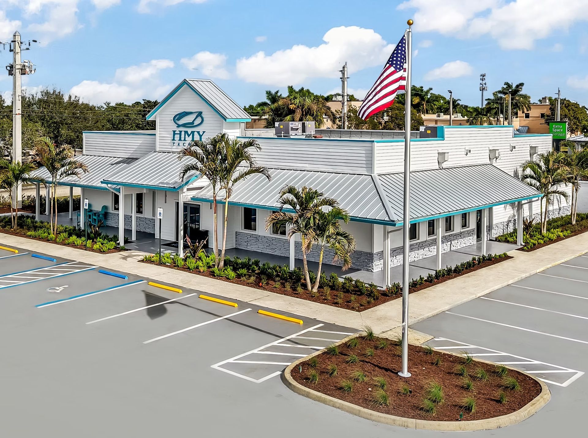 Aerial view of a white HYPER building with teal roof, palm trees, and a large American flag by a parking lot