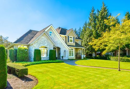 Suburban house with stone facade, manicured lawn, and tall trees under a bright blue sky