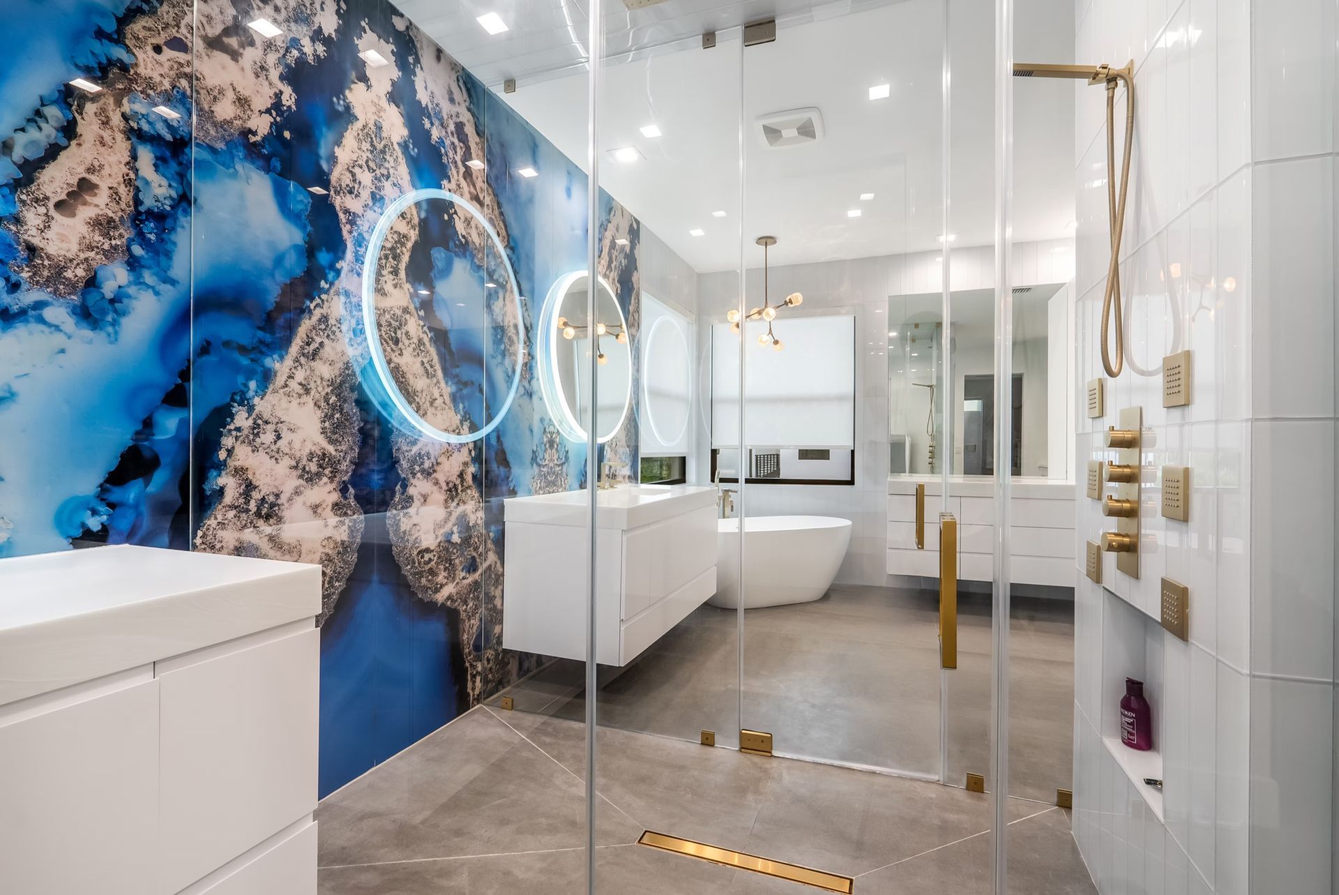 Modern bathroom with blue marble wall, round lit mirrors, glass shower, and gold fixtures