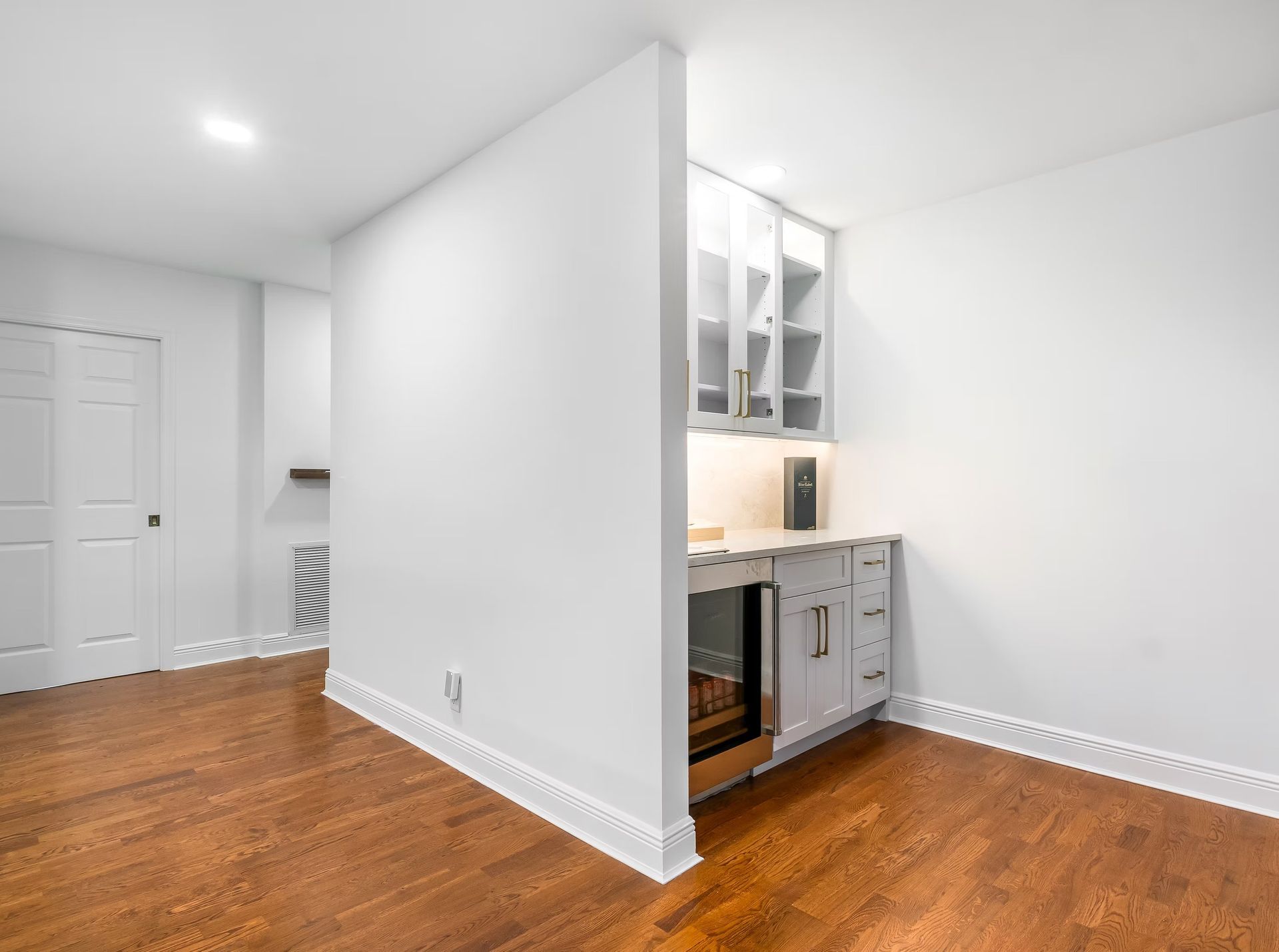 Empty modern room with hardwood floors, white walls, and a small kitchenette alcove.