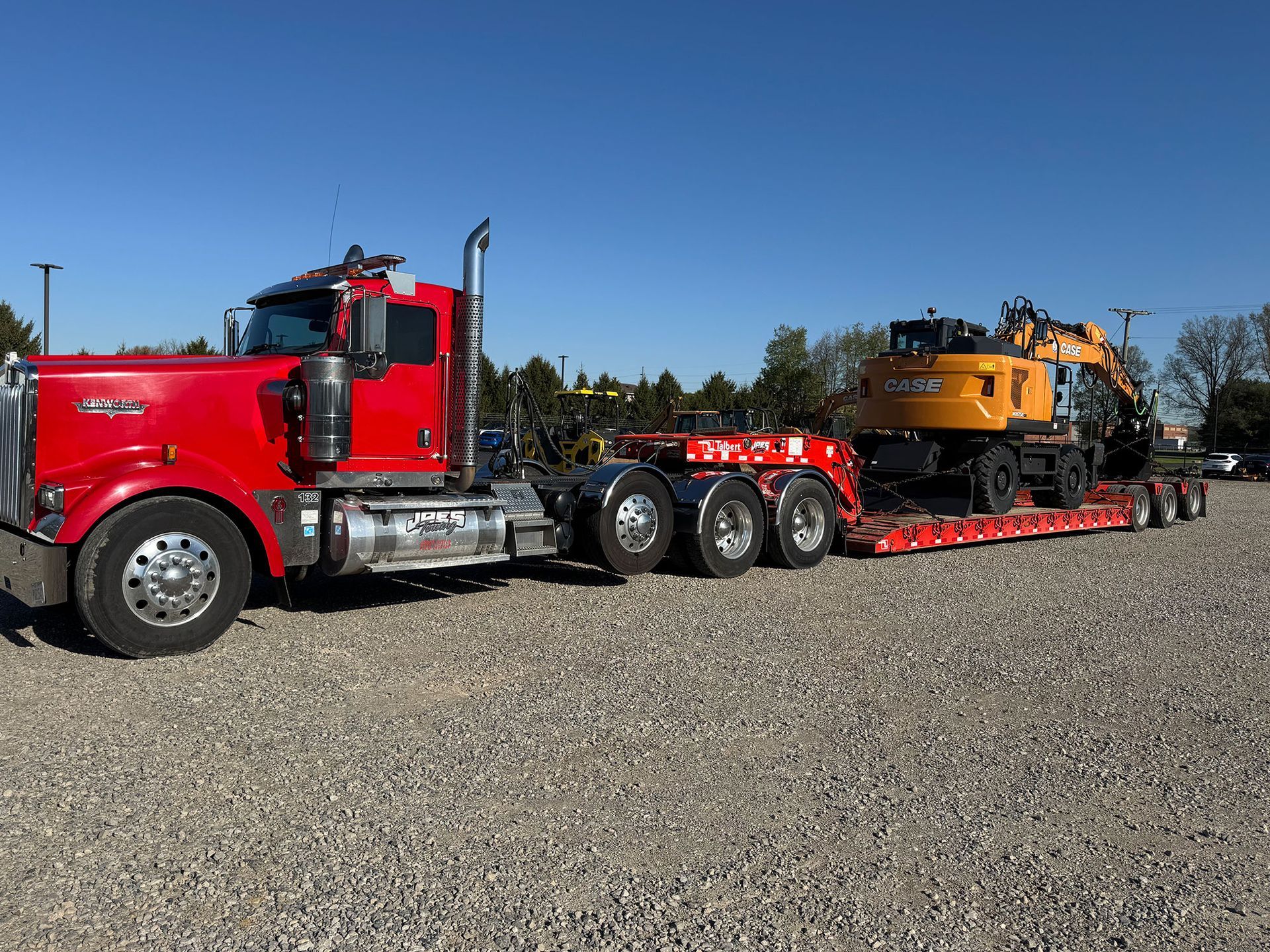 Red semi truck hauling an orange heavy equipment machine on a long red trailer in a gravel lot