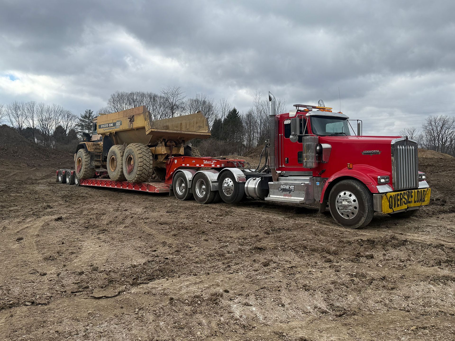 Red semi-truck hauling heavy equipment on a lowboy trailer in a muddy field under cloudy skies