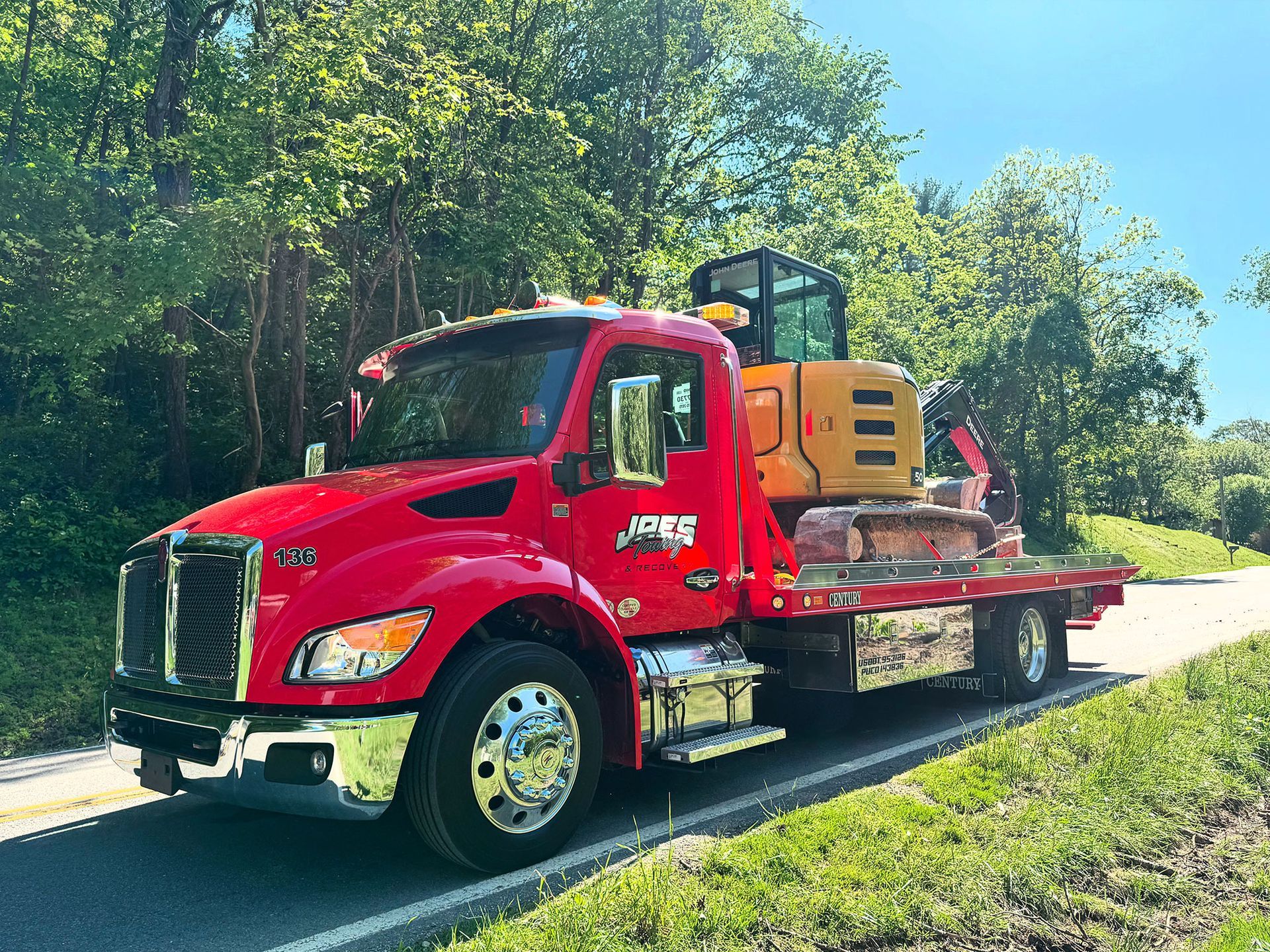 Red flatbed truck carrying a yellow machine on a sunny road beside trees and grass