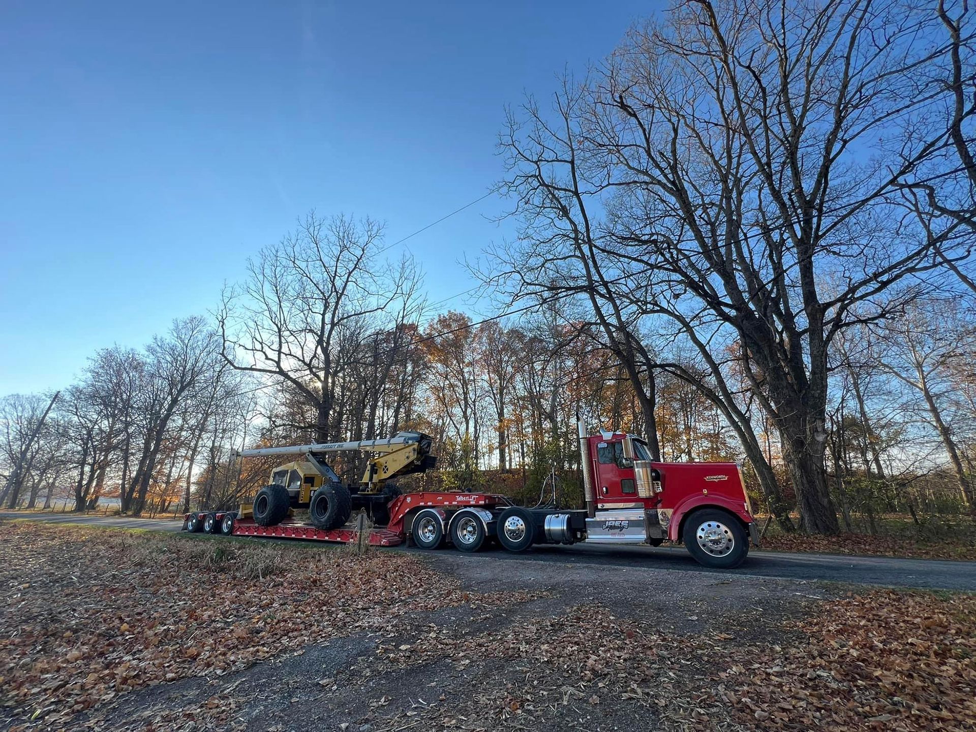 Red semi-truck hauling a yellow excavator on a flatbed trailer along a tree-lined road.