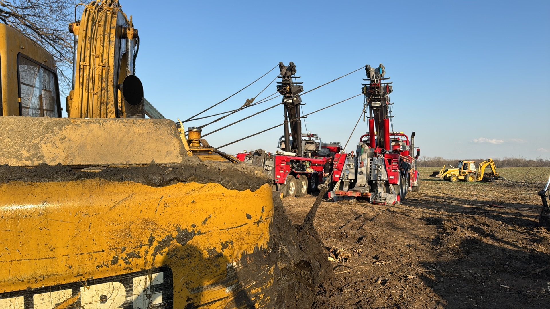Yellow excavator beside red drilling rigs in a muddy construction site under a clear sky