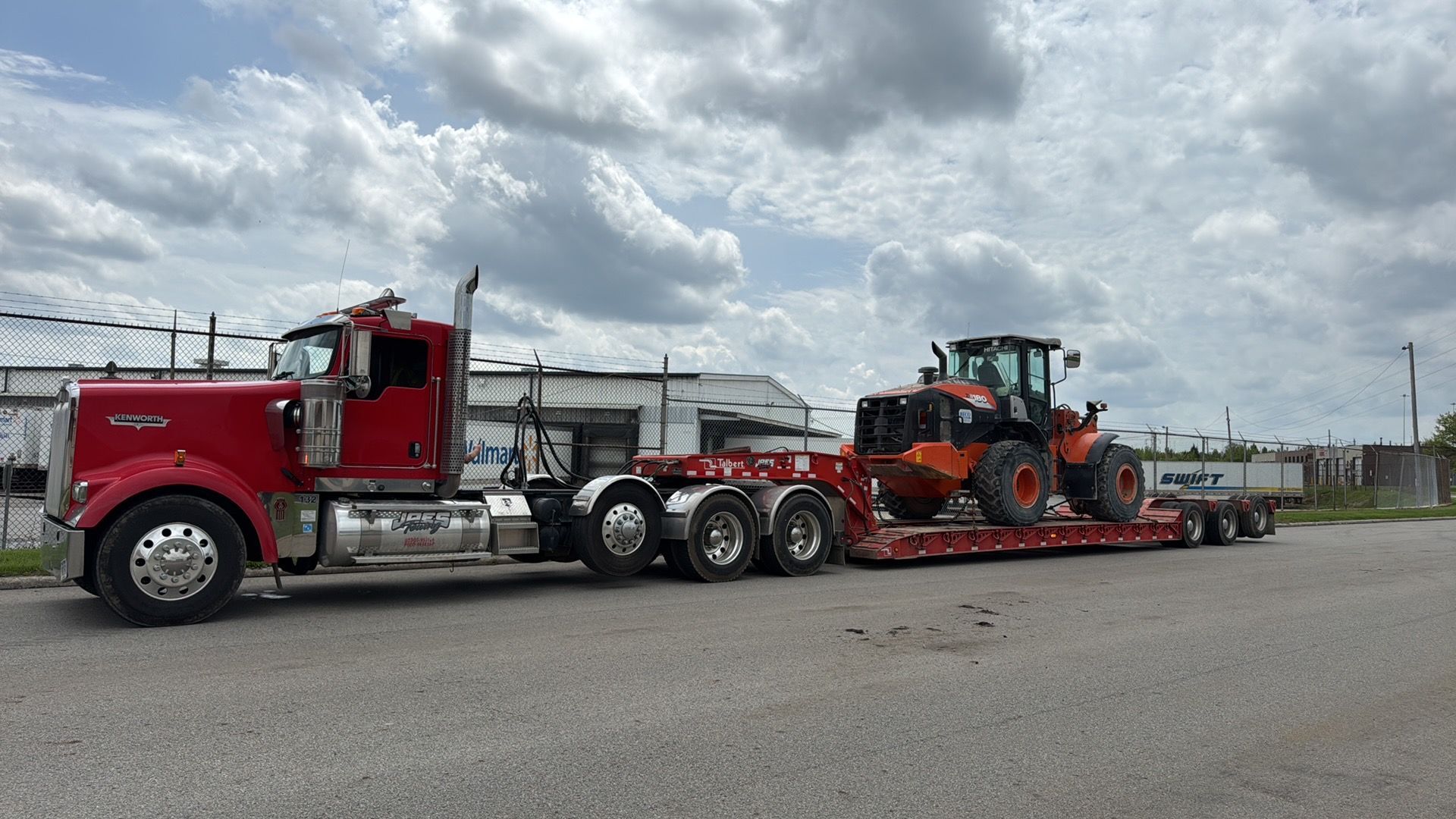 Red semi-truck hauling an orange tractor on a lowboy trailer in an industrial lot