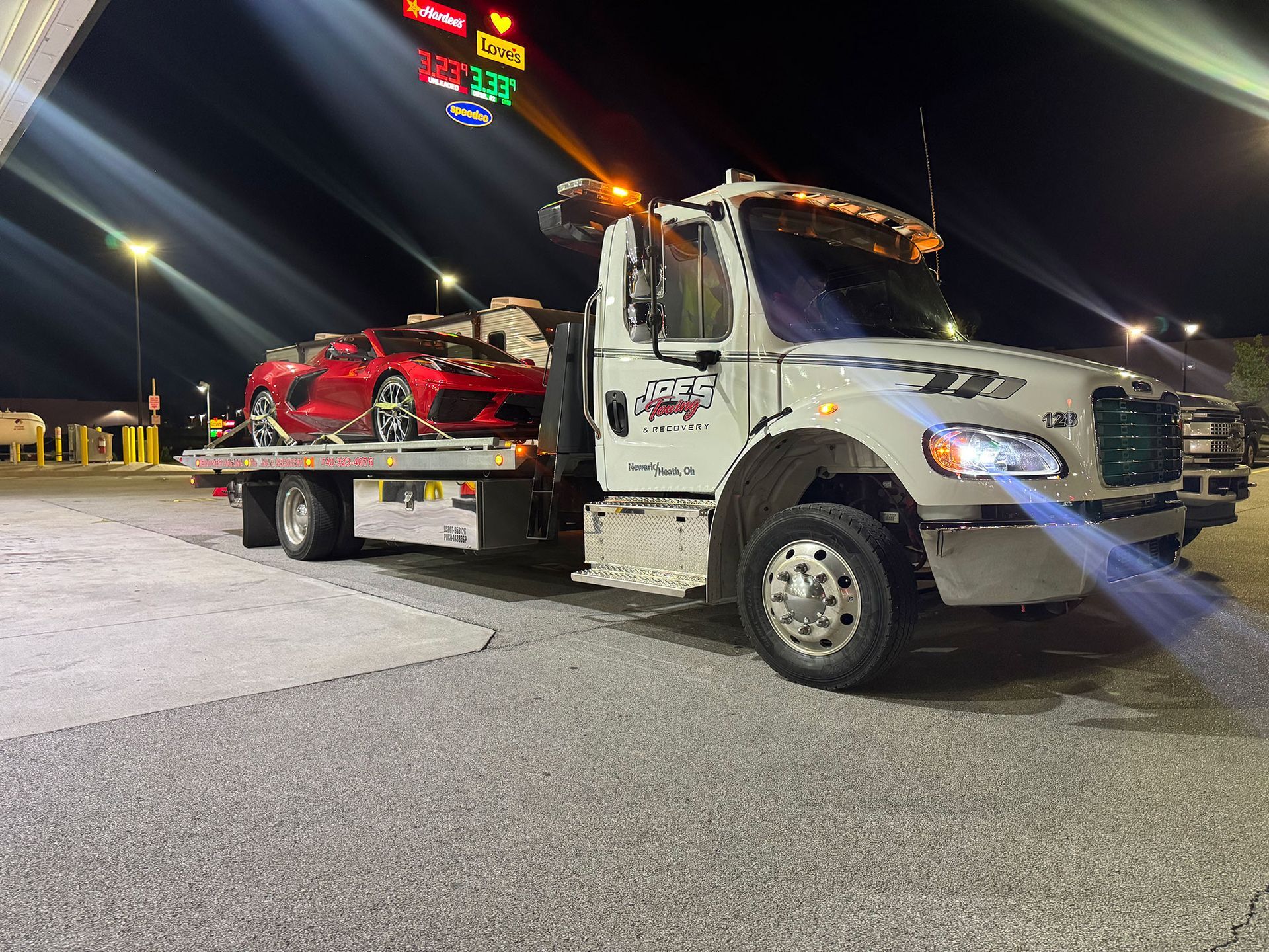 Tow truck carrying a damaged red sports car at night in a lit parking lot