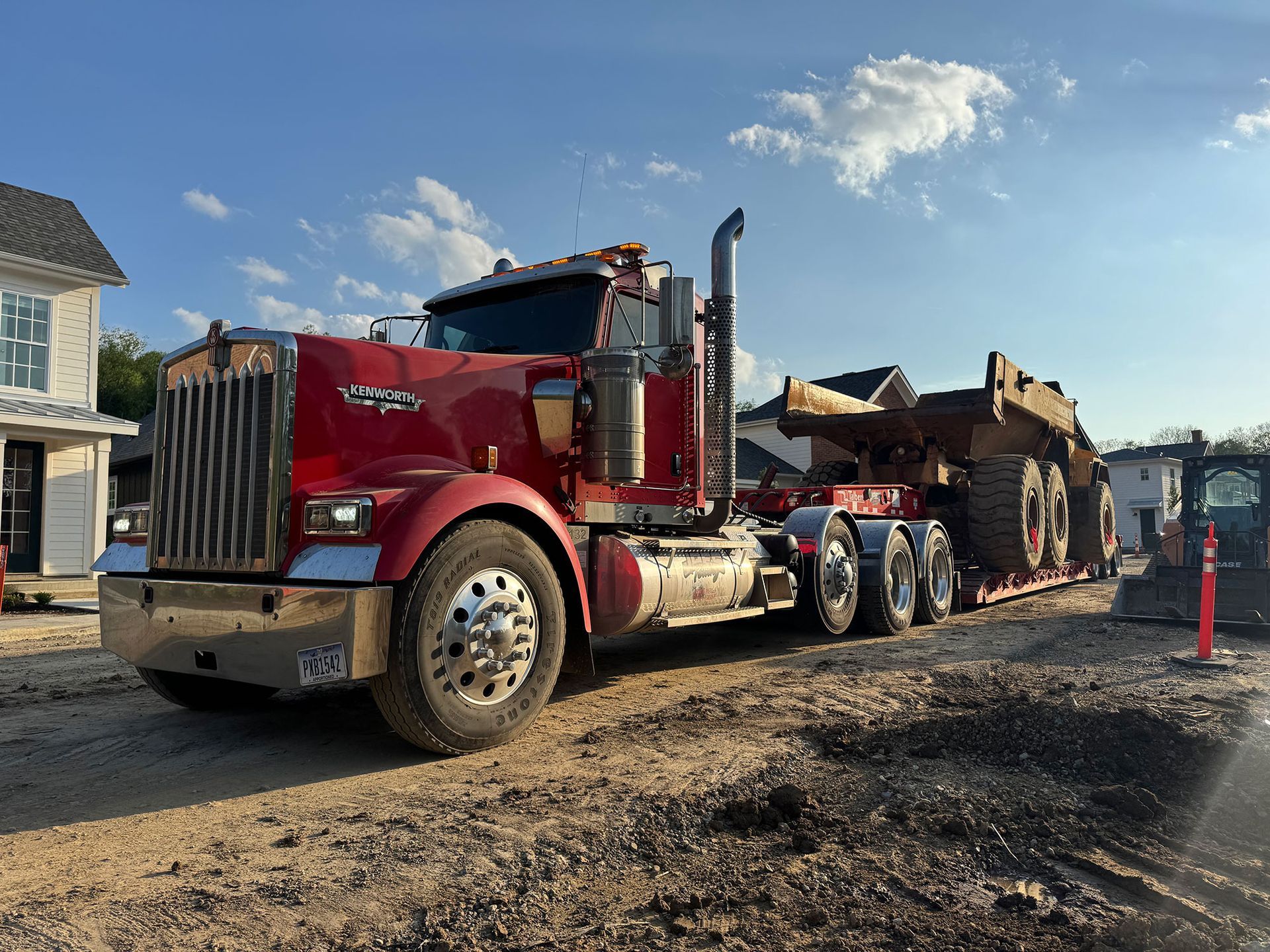 Red dump truck hauling a large load on a gravel lot, with construction cones and a house nearby