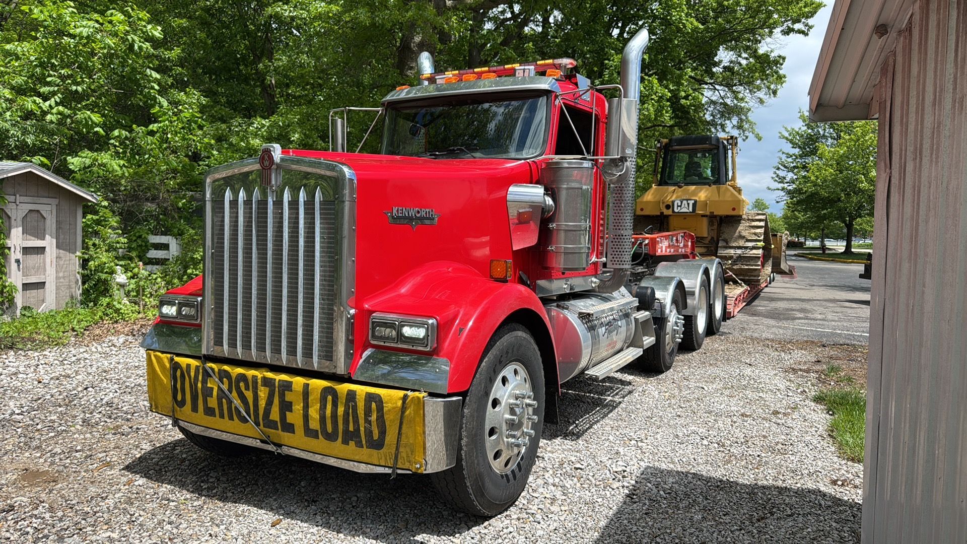 Red semi truck hauling a wide load on a trailer along a gravel driveway, surrounded by trees.