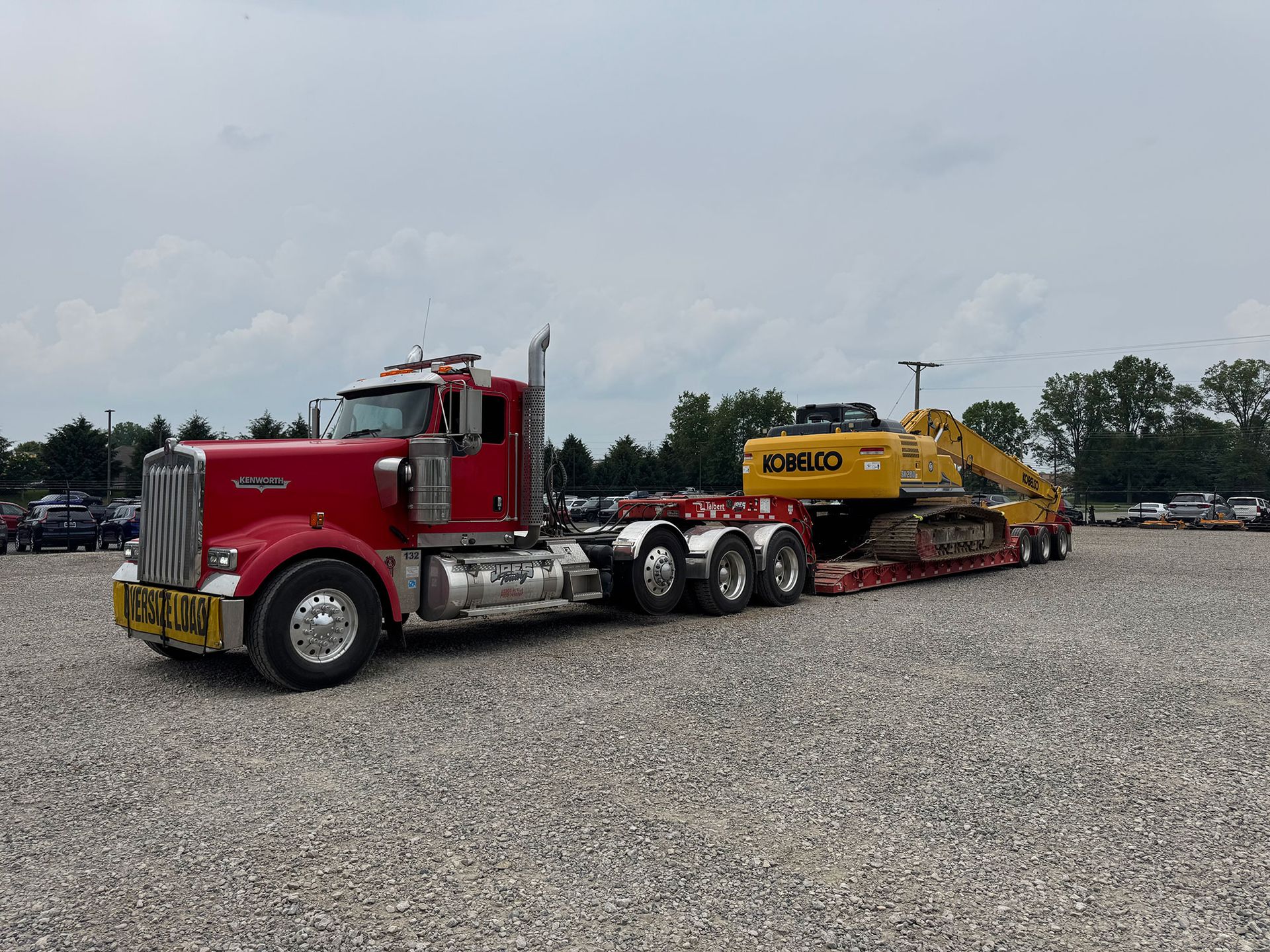 Red semi-truck hauling a yellow heavy machine on a flatbed in a gravel lot