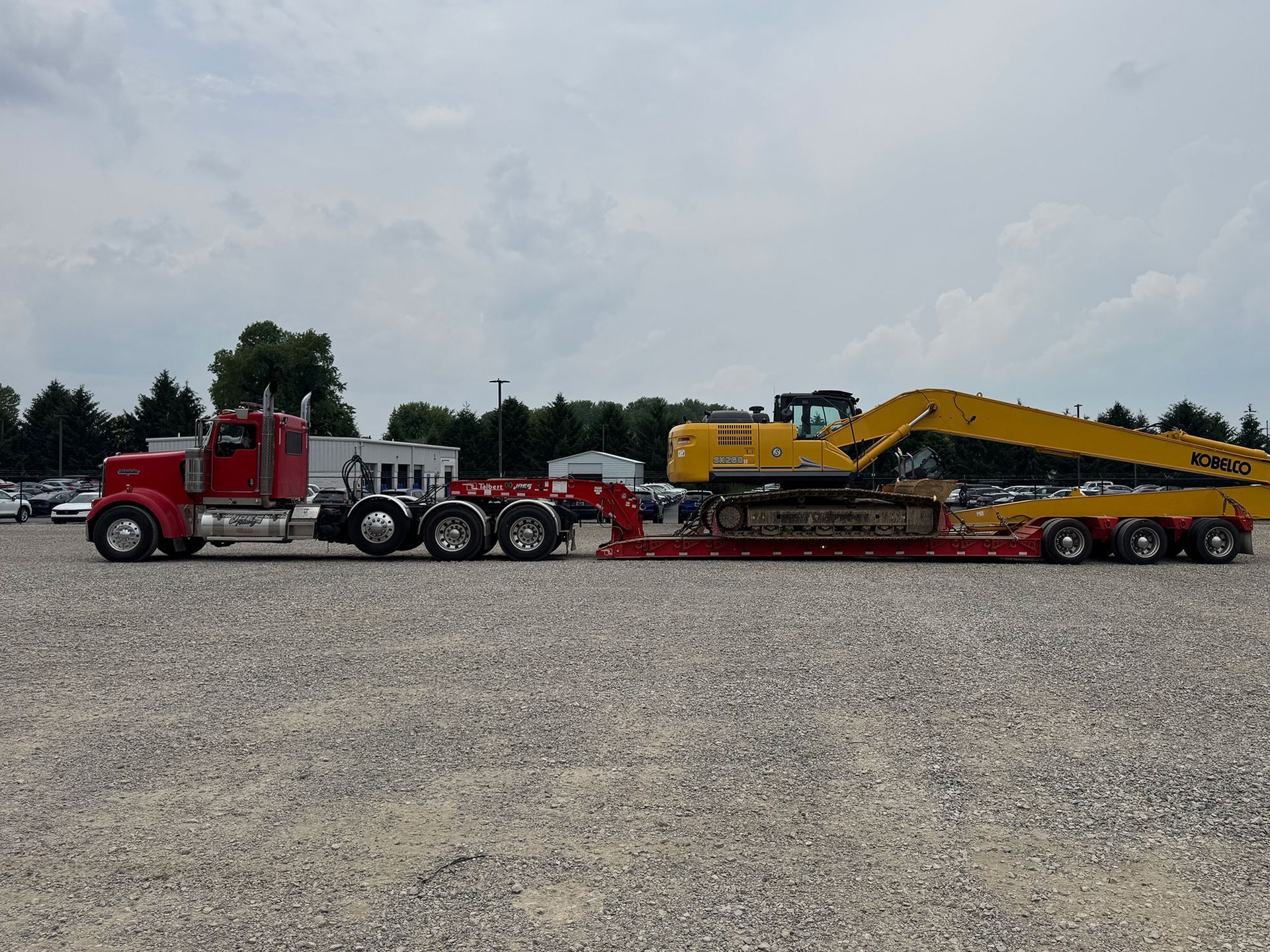 Red semi truck hauling a yellow excavator on a long flatbed trailer in a gravel lot