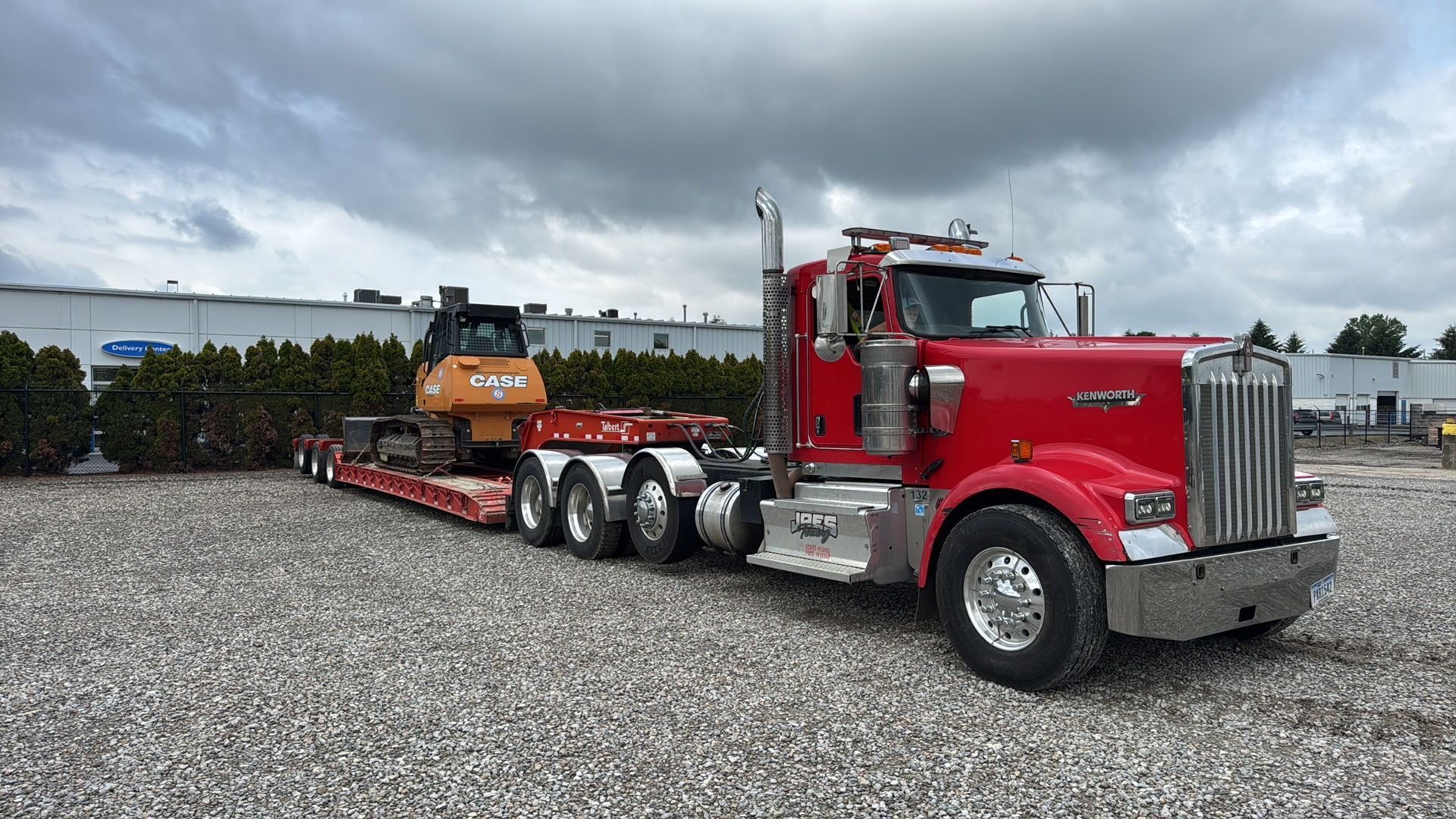 Red semi truck hauling an orange excavator on a long lowboy trailer in a gravel lot