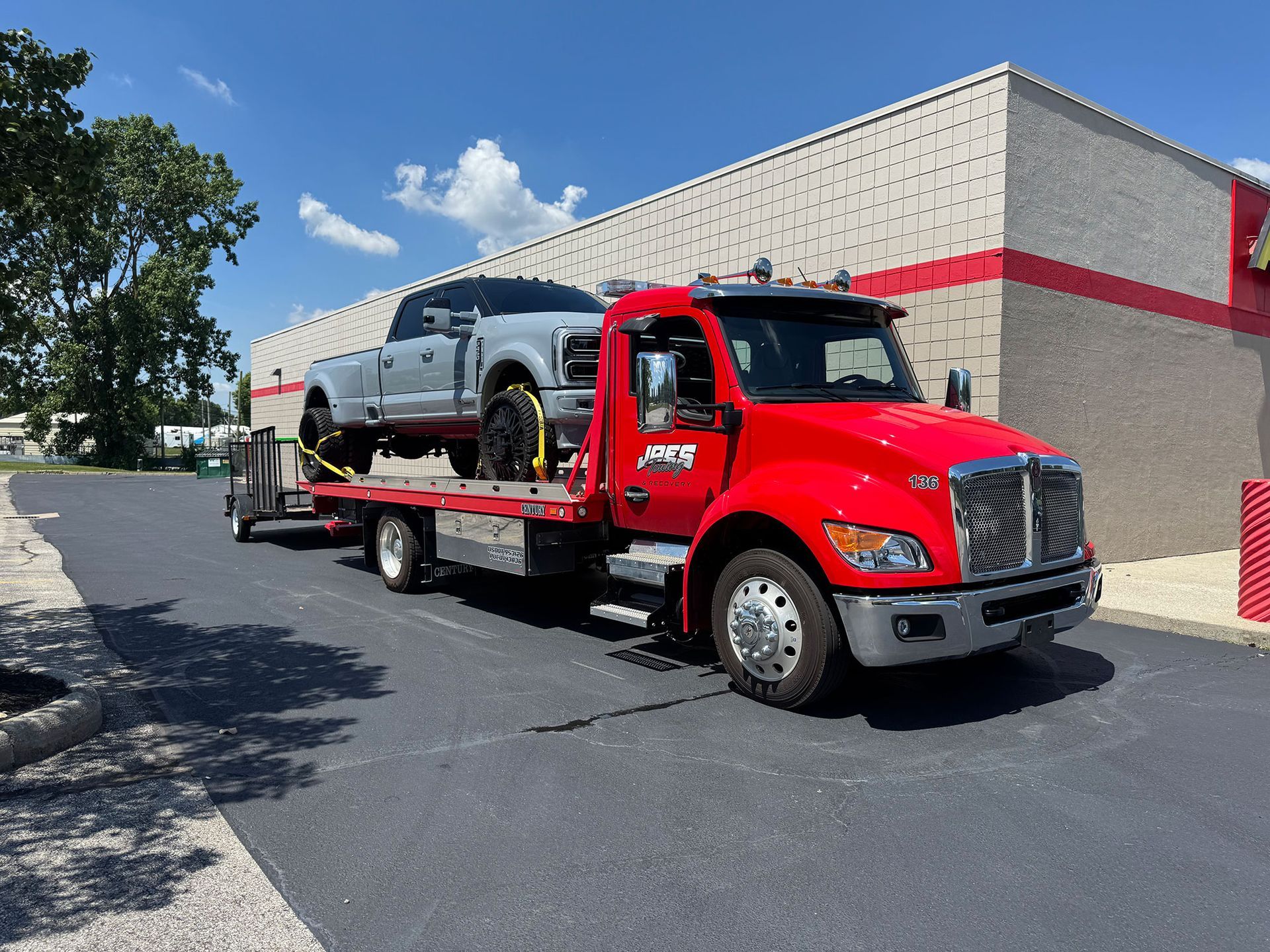 Red tow truck carrying a silver pickup outside a building on a sunny day