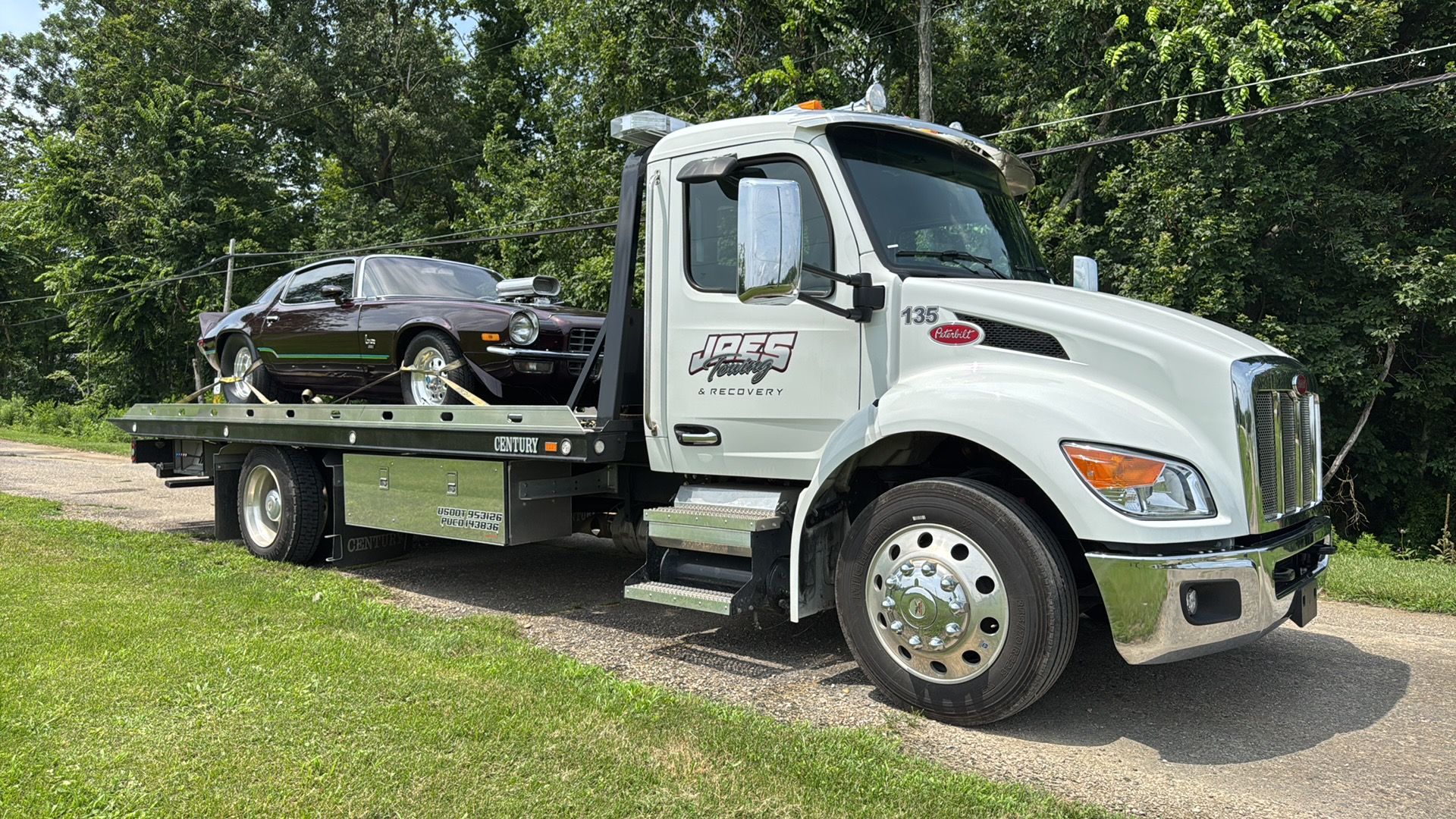 White flatbed tow truck carrying a dark sedan on a road beside grass and trees
