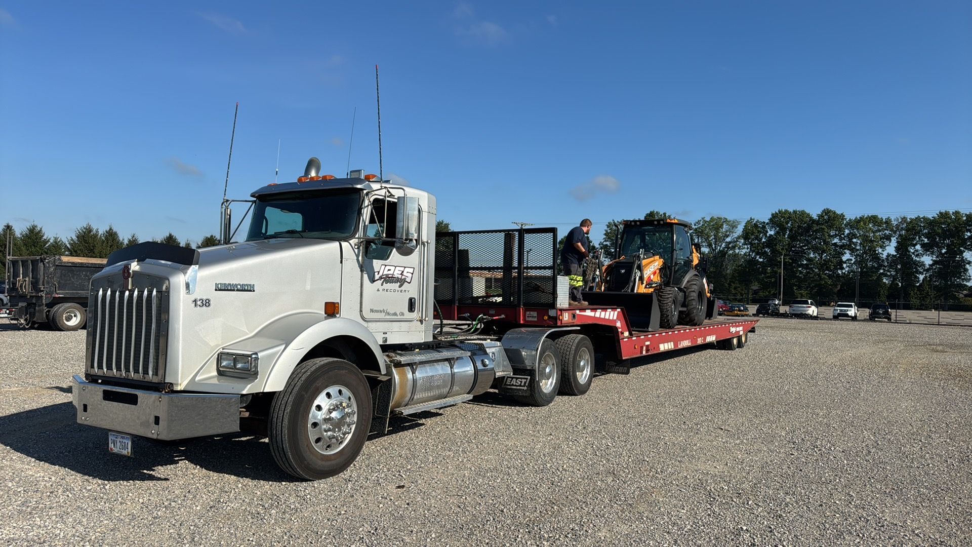 White semi truck hauling a red flatbed trailer with a forklift on a gravel lot under a clear blue sky