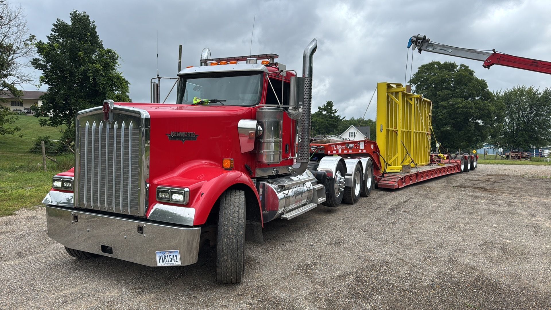 Red semi truck hauling a yellow industrial load on a flatbed in a gravel lot under cloudy skies