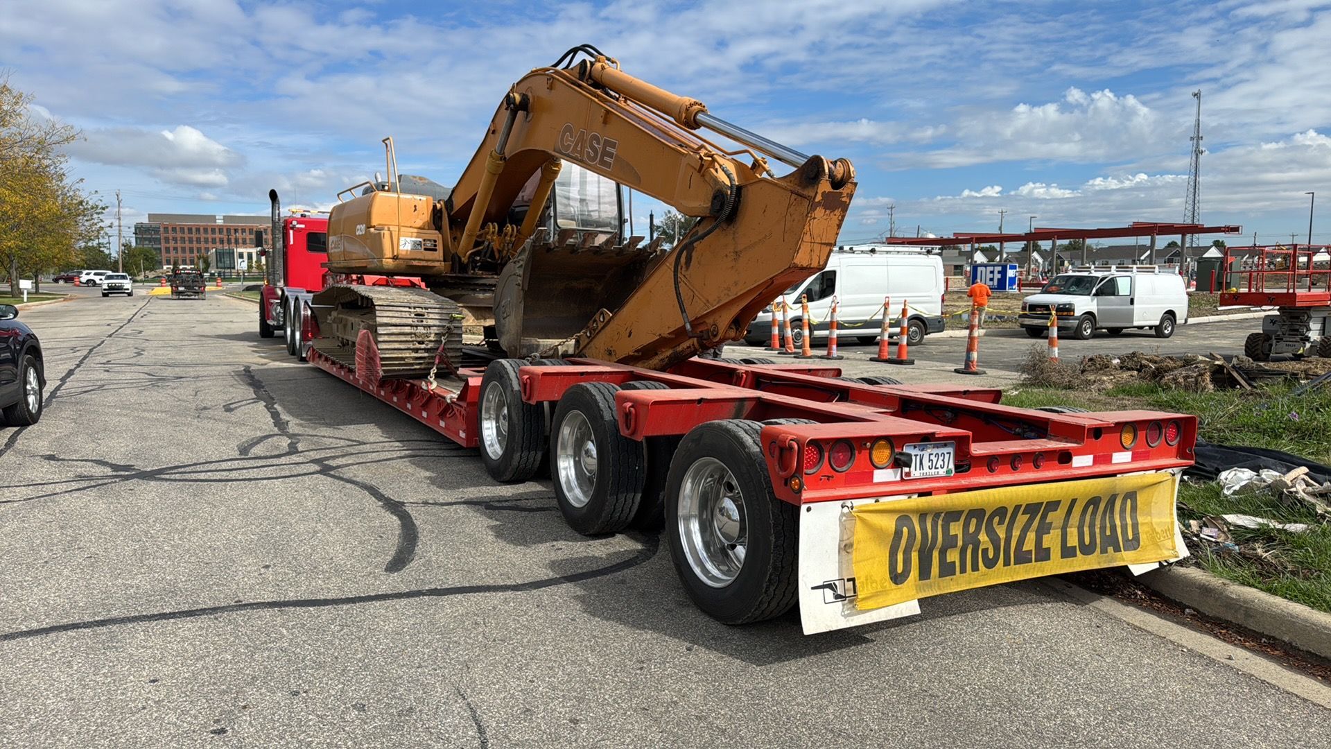 Excavator loaded on a red flatbed trailer on a road, with yellow oversize-load banner at rear.