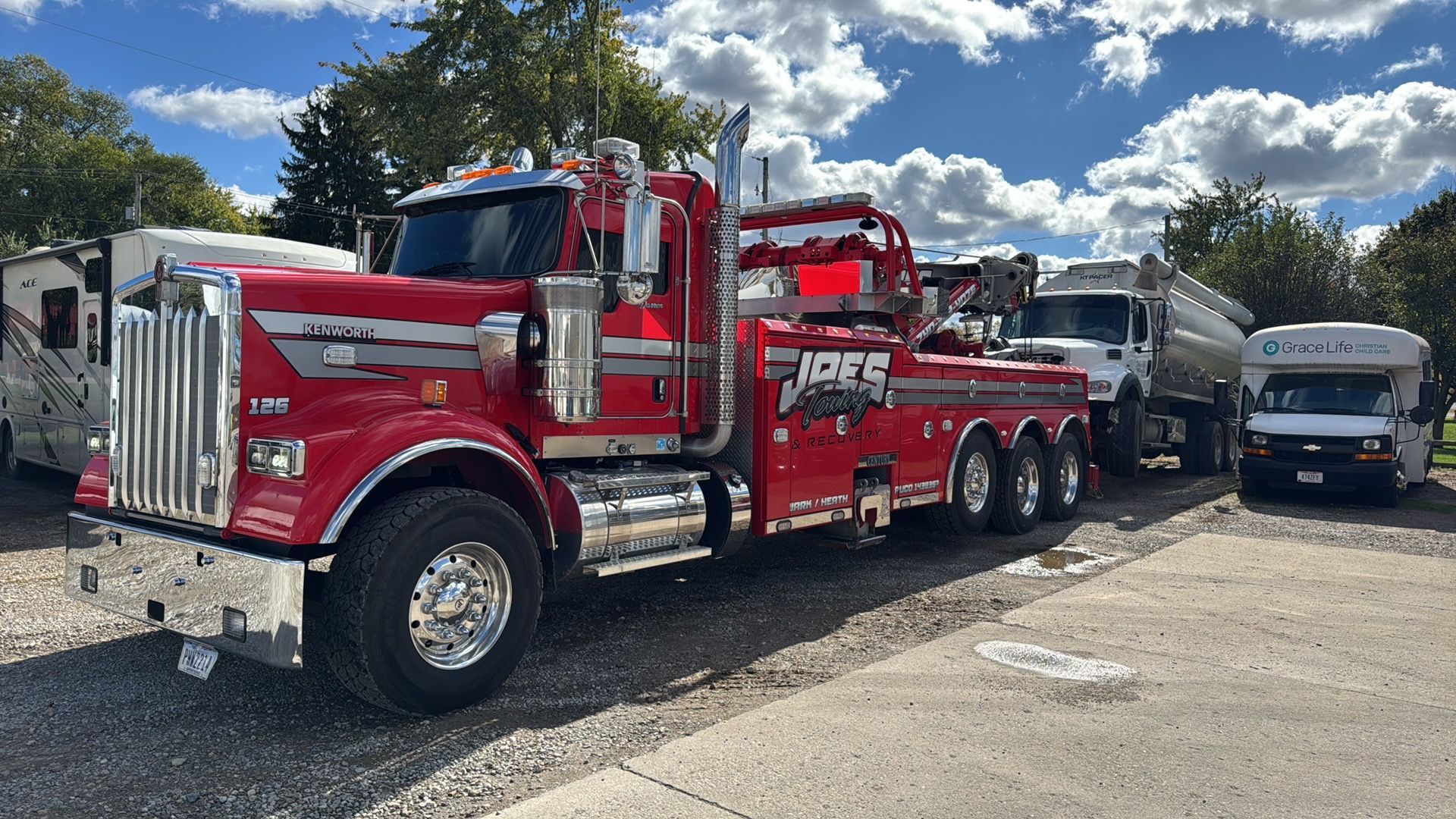 Red and silver tow truck parked on a gravel lot beside a white van under a blue sky