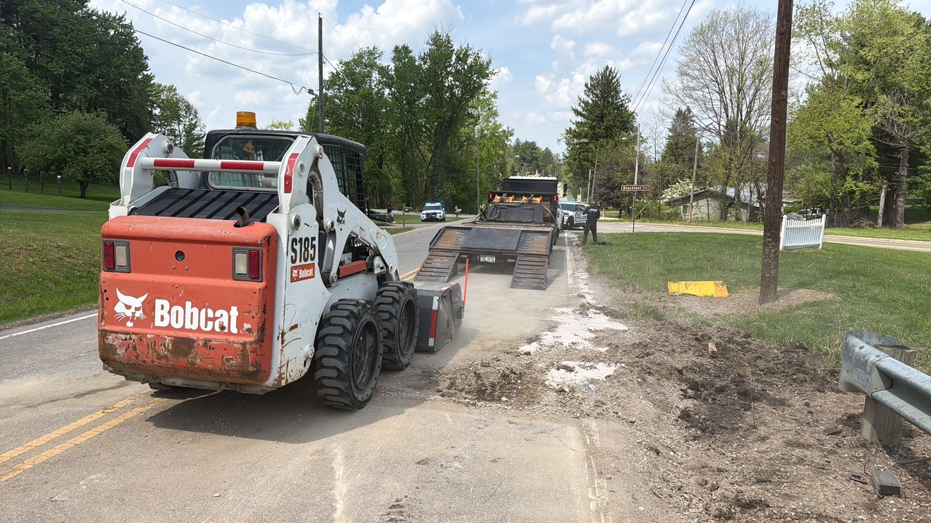 Bobcat skid steer grading a gravel road with a dump truck nearby on a rural street