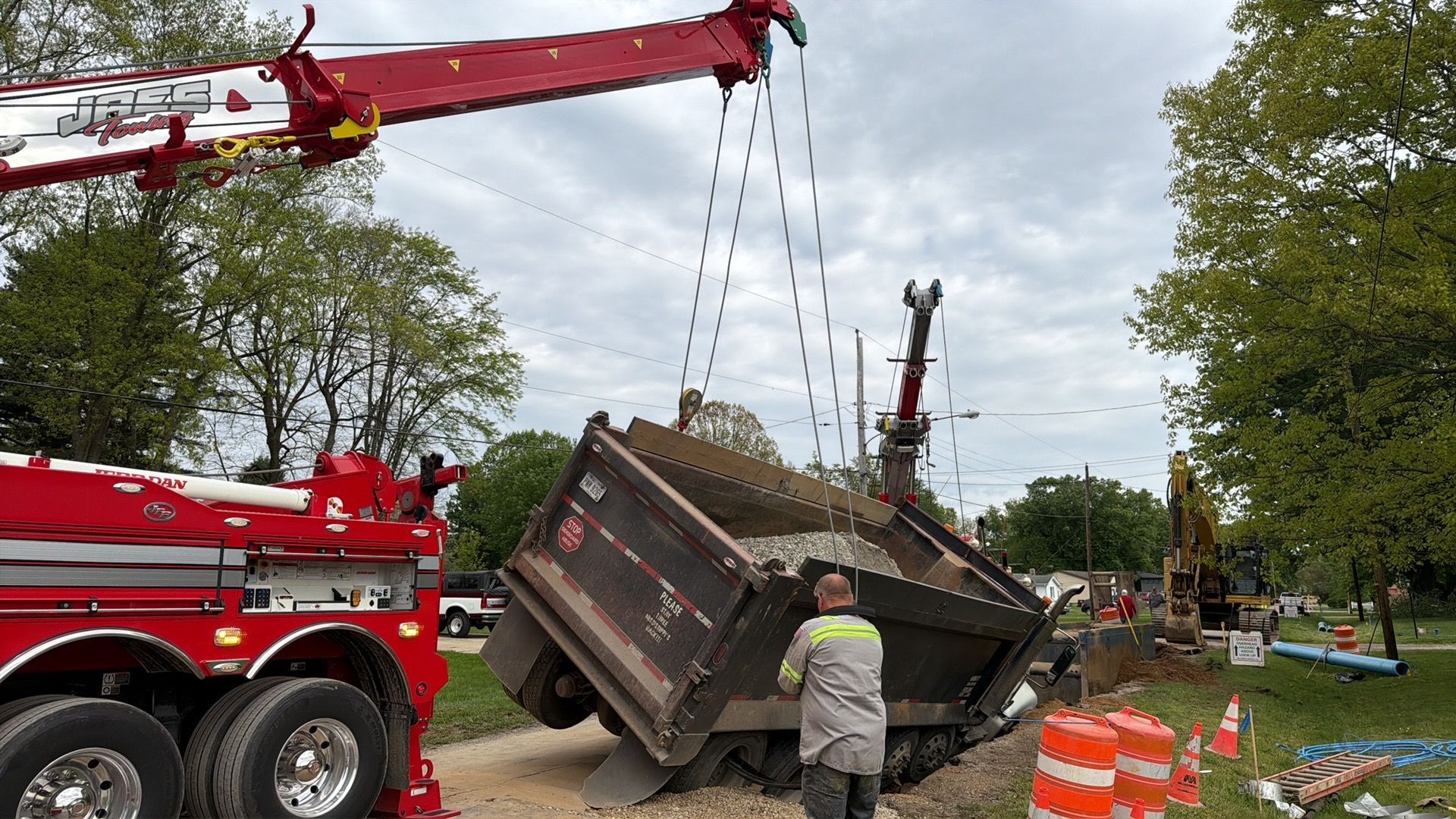 Tow truck lifting a tipped dump trailer on a roadside, with a worker standing nearby.