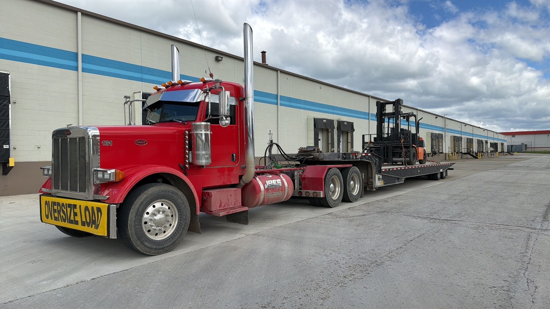 Red semi-truck hauling a flatbed trailer loaded with industrial equipment beside a warehouse