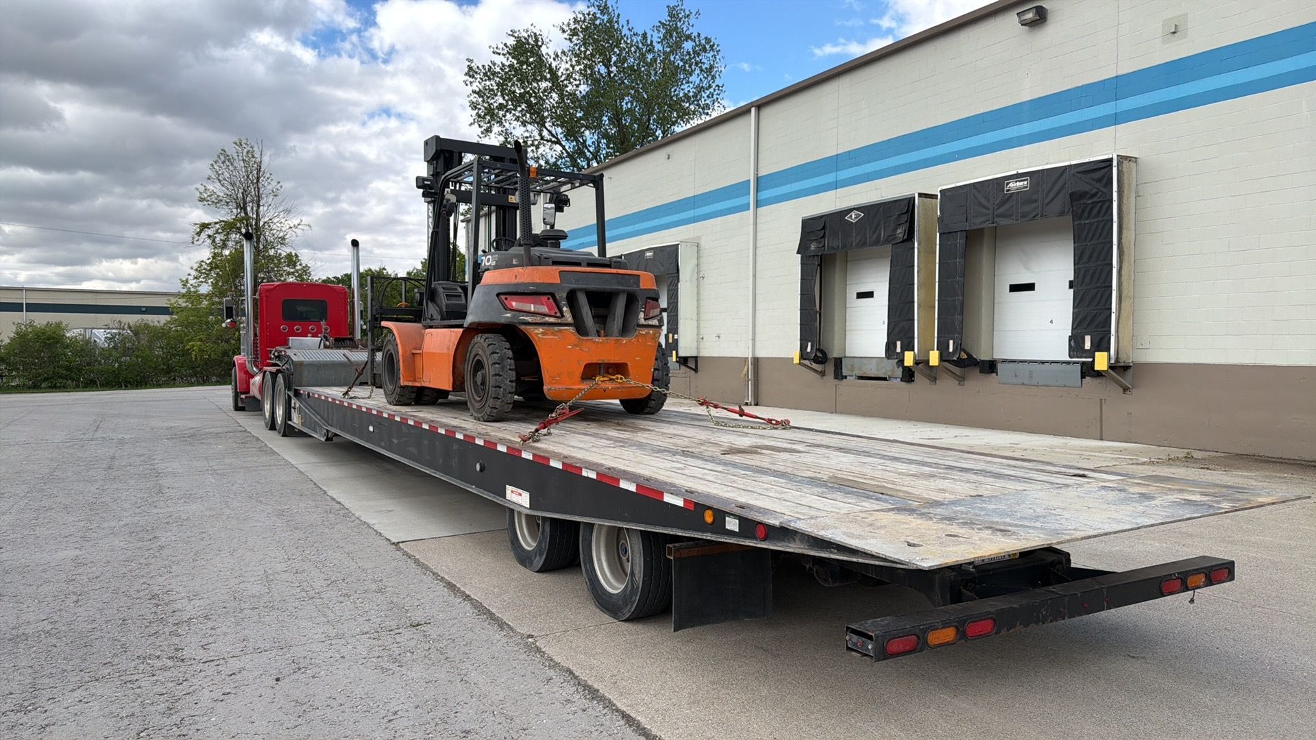Orange forklift on a flatbed trailer beside a warehouse loading dock
